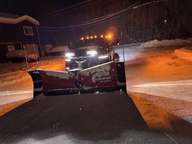 A snow-covered street at night with a snowplow truck pushing snow. Houses and parked cars are visible in the background.