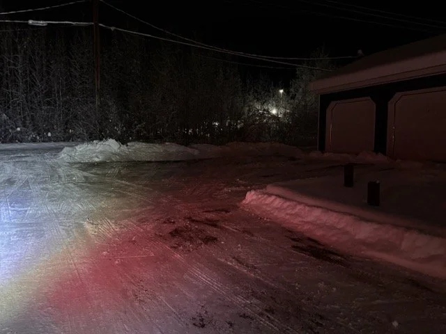 Snow-covered driveway and garage at night with faint streetlights in the background.