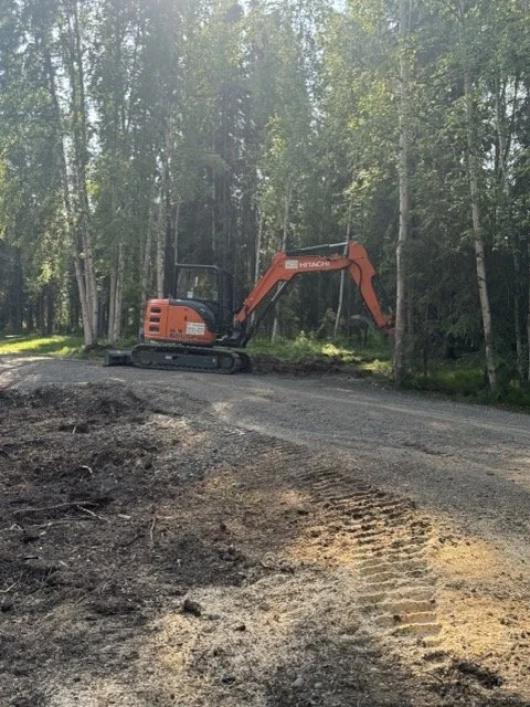 An orange excavator working on a dirt path near a wooded area with green trees.