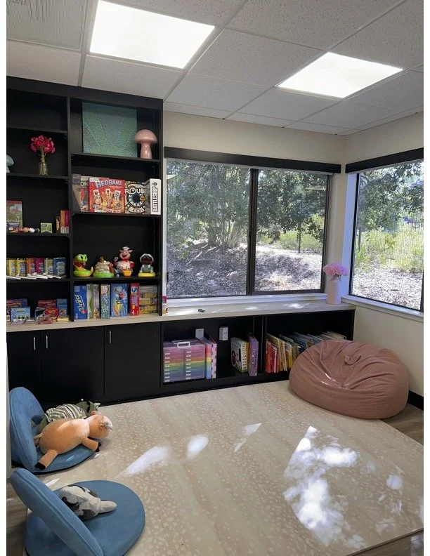 A cozy playroom with large windows showing trees outside. It has a black bookshelf filled with board games, books, and toys, and a pink vase with flowers on top. There are a blue chair with a stuffed animal, a pink bean bag, and a beige rug with white patterns on the floor.