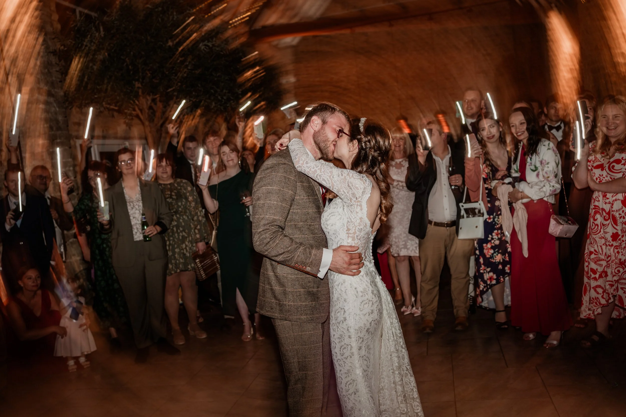 Bride and groom sharing a kiss at their wedding reception while guests watch and celebrate in the background, under a brick arch ceiling with warm lighting.