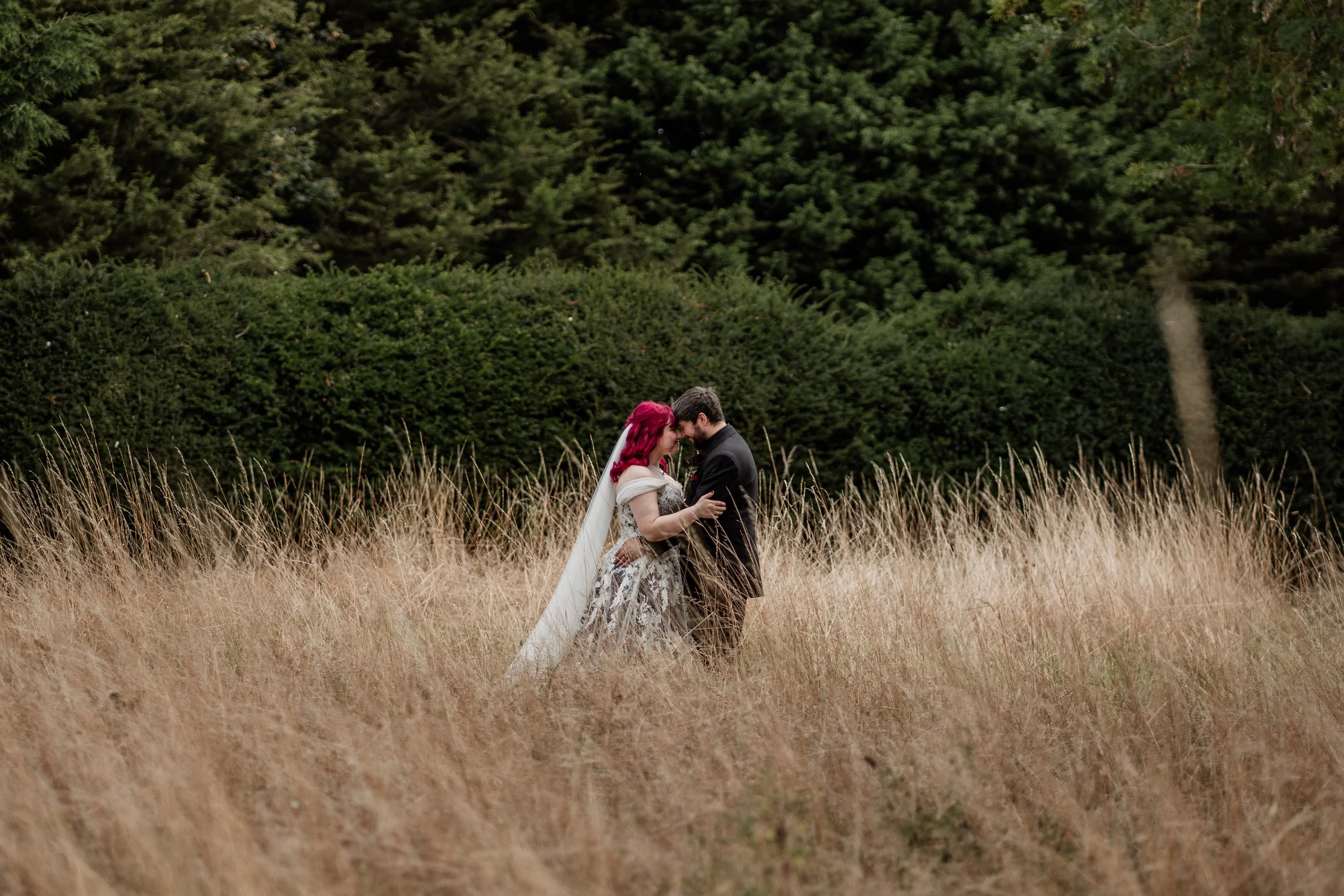 A couple dressed in wedding attire standing close together in a field of tall, golden grass, with a background of dense green trees.