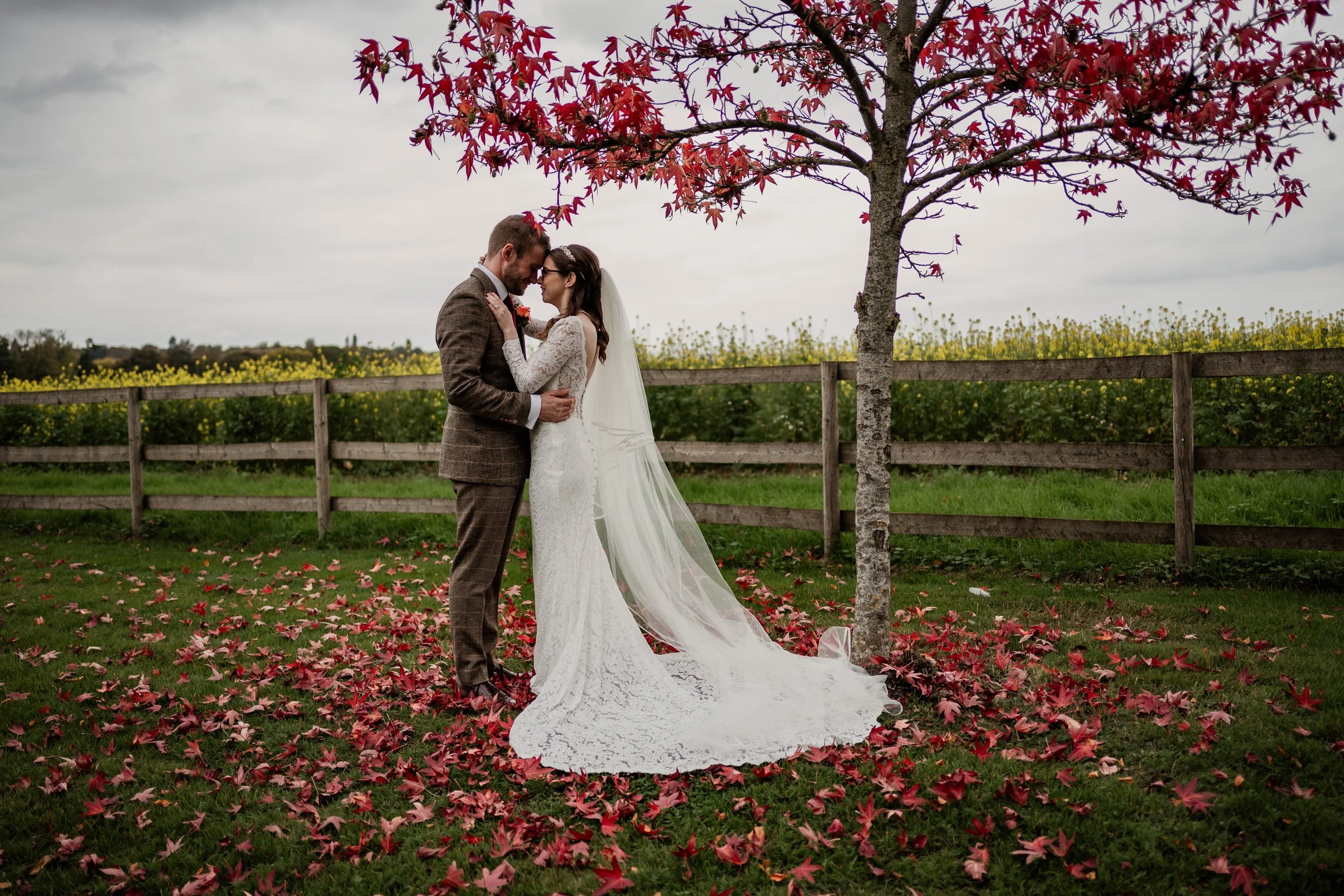 A bride and groom standing close together under a pink-leafed tree on a grassy field, with fall leaves on the ground and a wooden fence in the background, sharing an intimate moment on their wedding day.