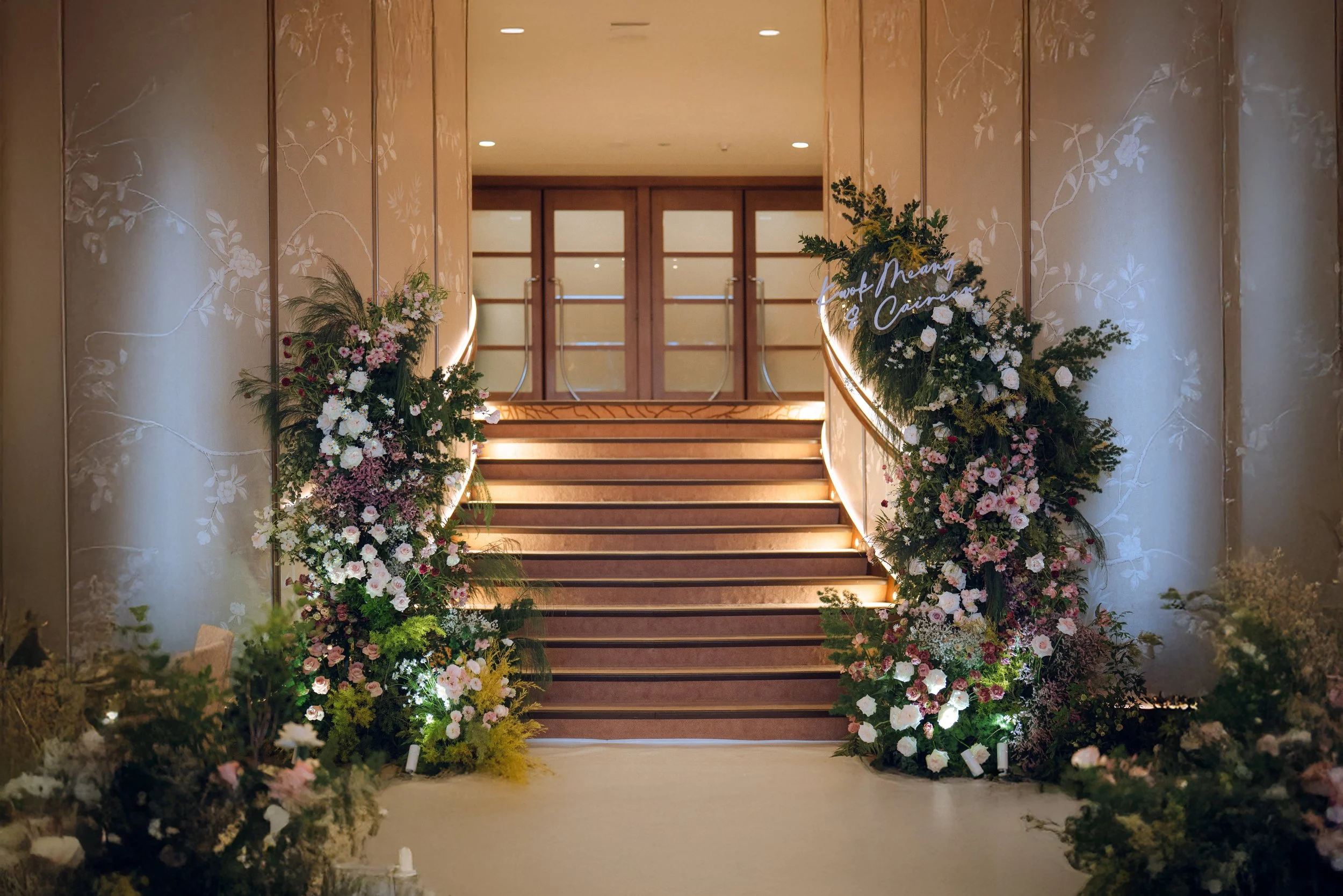 Indoor wedding ceremony setup with white flowers, green plants, and wooden chairs arranged on both sides of the aisle in front of an arch with hanging lanterns.