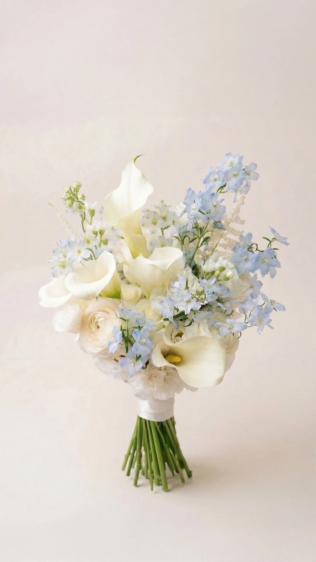 A bouquet of white and pale yellow flowers, including calla lilies, roses, and delphiniums, arranged in a white wrap with green stems visible, set against a plain, light background.