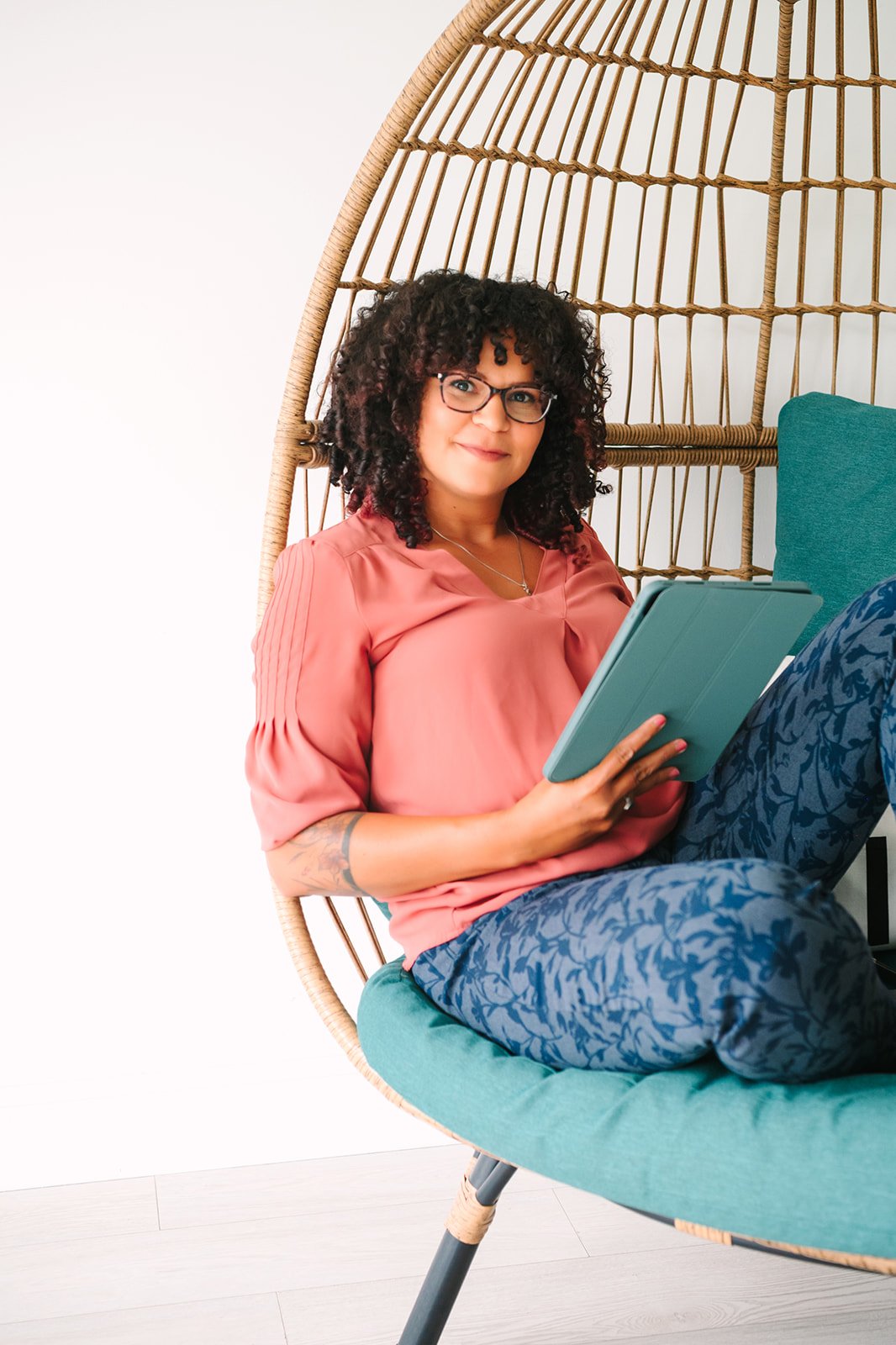 Latreash with glasses, and tattoos, sitting in a hanging wicker chair with teal cushion, holding a tablet, wearing a pink blouse and patterned blue pants.