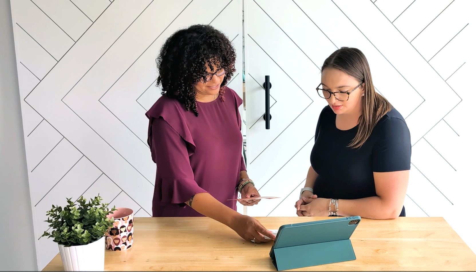 Overhead view of a person working at a desk with a laptop, a monitor displaying graphic design software, a pink keyboard, a black mouse, a notebook with a pen, a small plant, and a cup with pens. The desk is next to a window with a purple curtain.