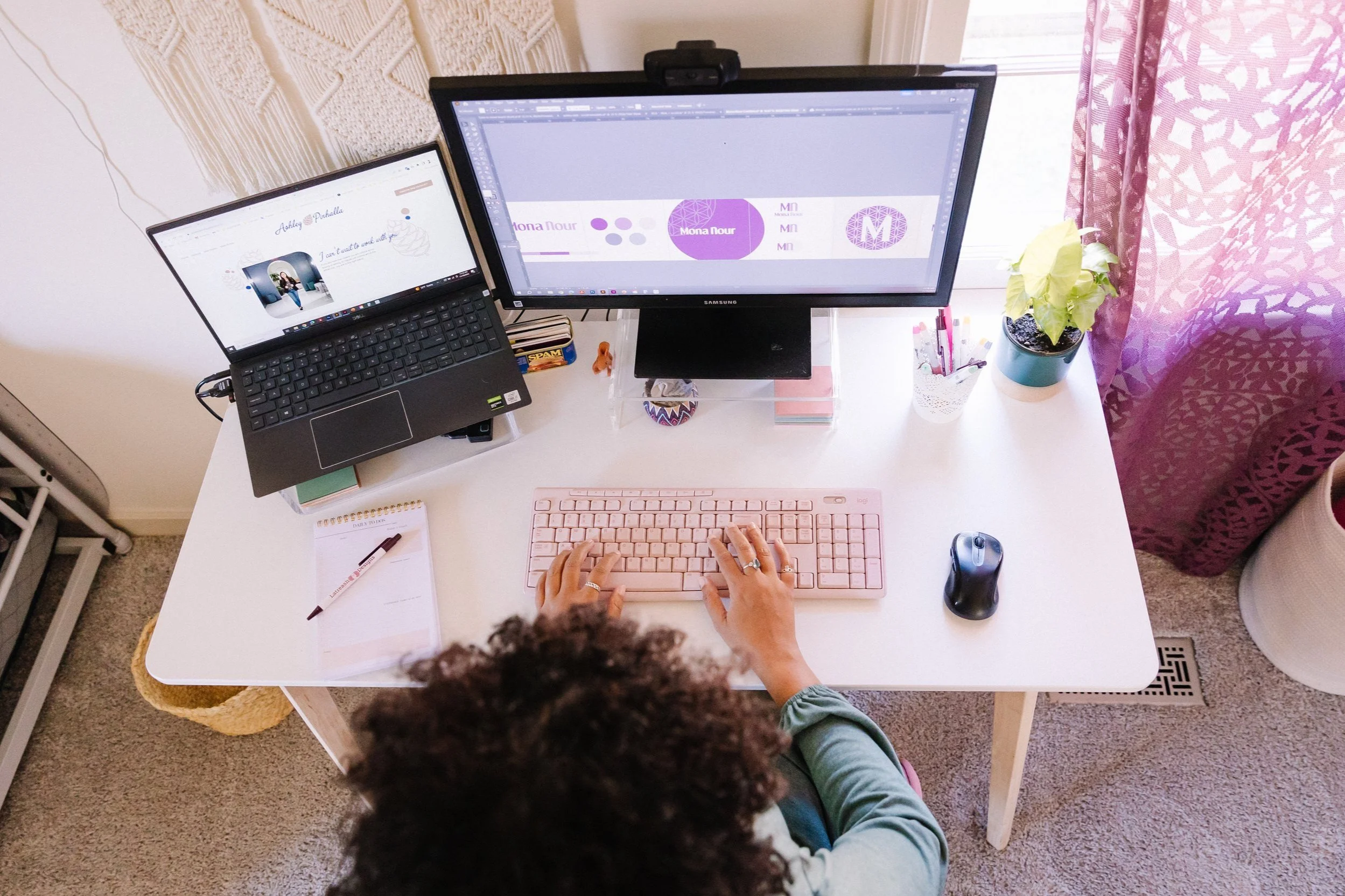 Overhead view of a person working at a desk with a laptop, a monitor displaying graphic design software, a pink keyboard, a black mouse, a notebook with a pen, a small plant, and a cup with pens. The desk is next to a window with a purple curtain.