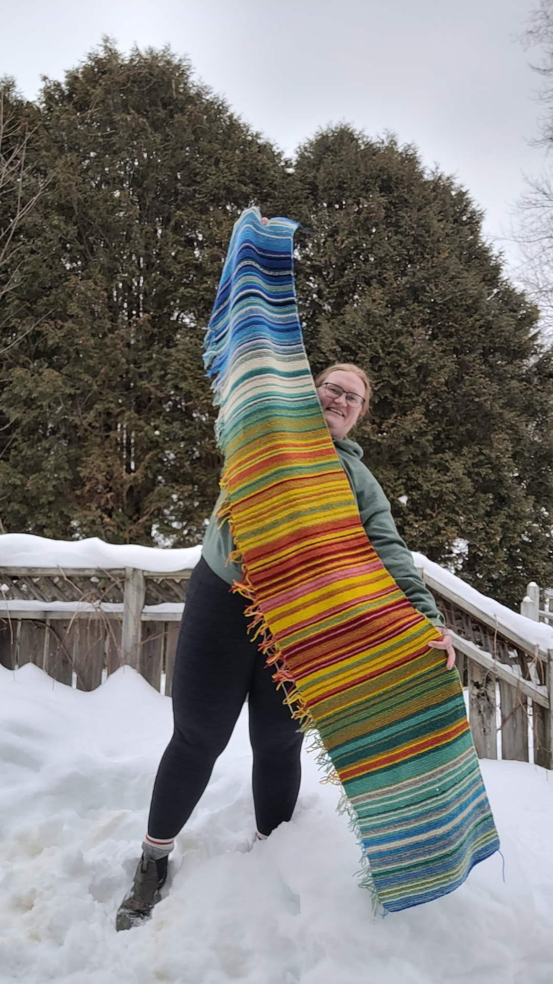A woman holding a temperature blanket section on a snow covered patio. There are trees in the background that are green and do not have snow.