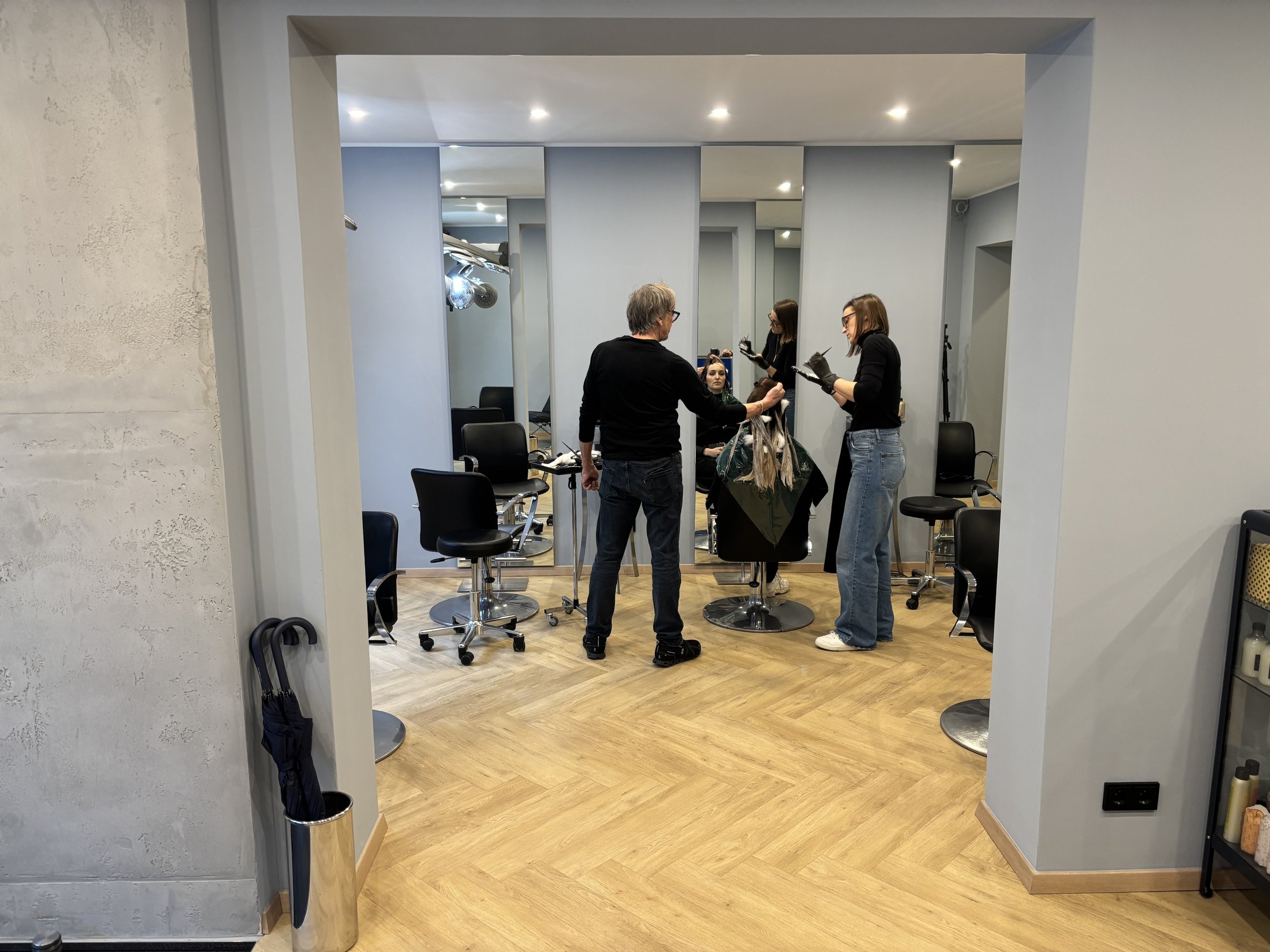 Ein Friseursalon mit zwei Friseurmeistern, die mit einem Kunden arbeiten. Der Kunde sitzt im Friseurstuhl, während die Friseurinnen die Haare in Abschnitte trennen und Farbe auftragen. Es sind mehrere Friseurstühle und Spiegel im Hintergrund.