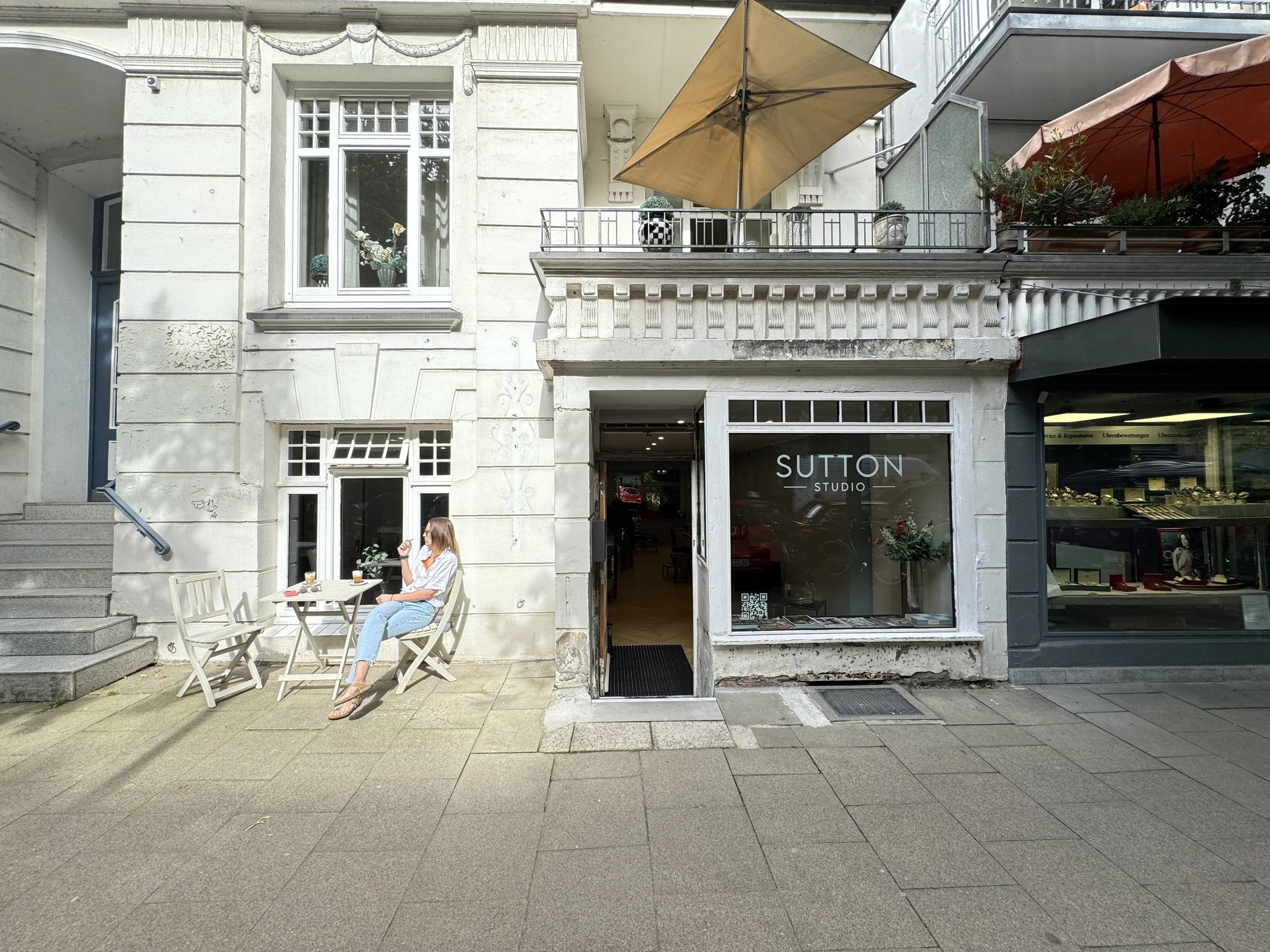 Frau sitzt im Außenbereich vor dem Salon "SUTTON STUDIO", trinkt Kaffee und liest ein Buch, in der Sonne, mit Getränken auf dem Tisch.