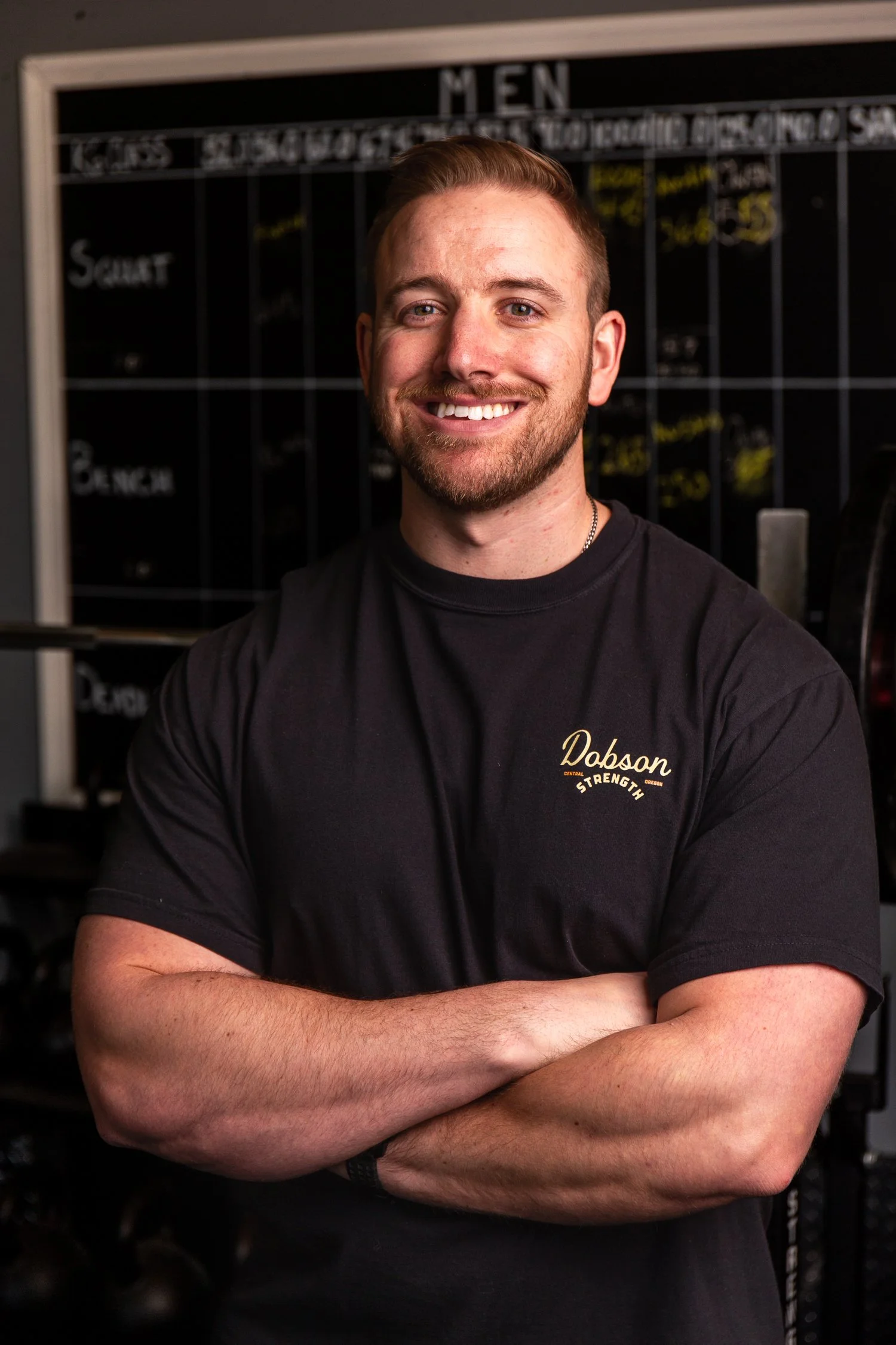 A smiling man with a beard and short hair, wearing a black t-shirt with 'Dobson Strength' written on it, standing with arms crossed in a gym with a blackboard in the background.
