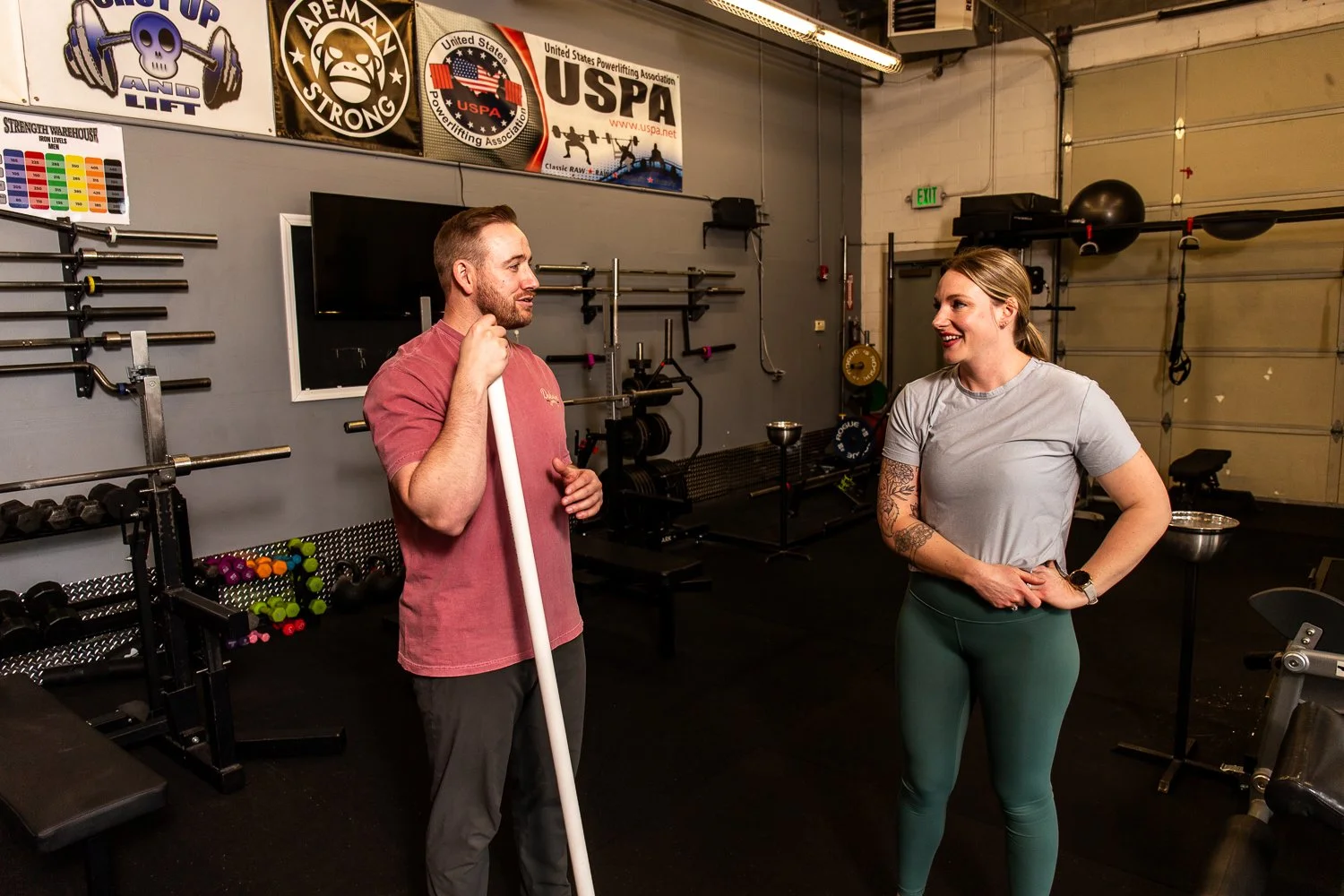 Two people talking in a gym, one man holding a white pipe and a woman smiling, fitness equipment, banners on the wall, and gym accessories in the background.