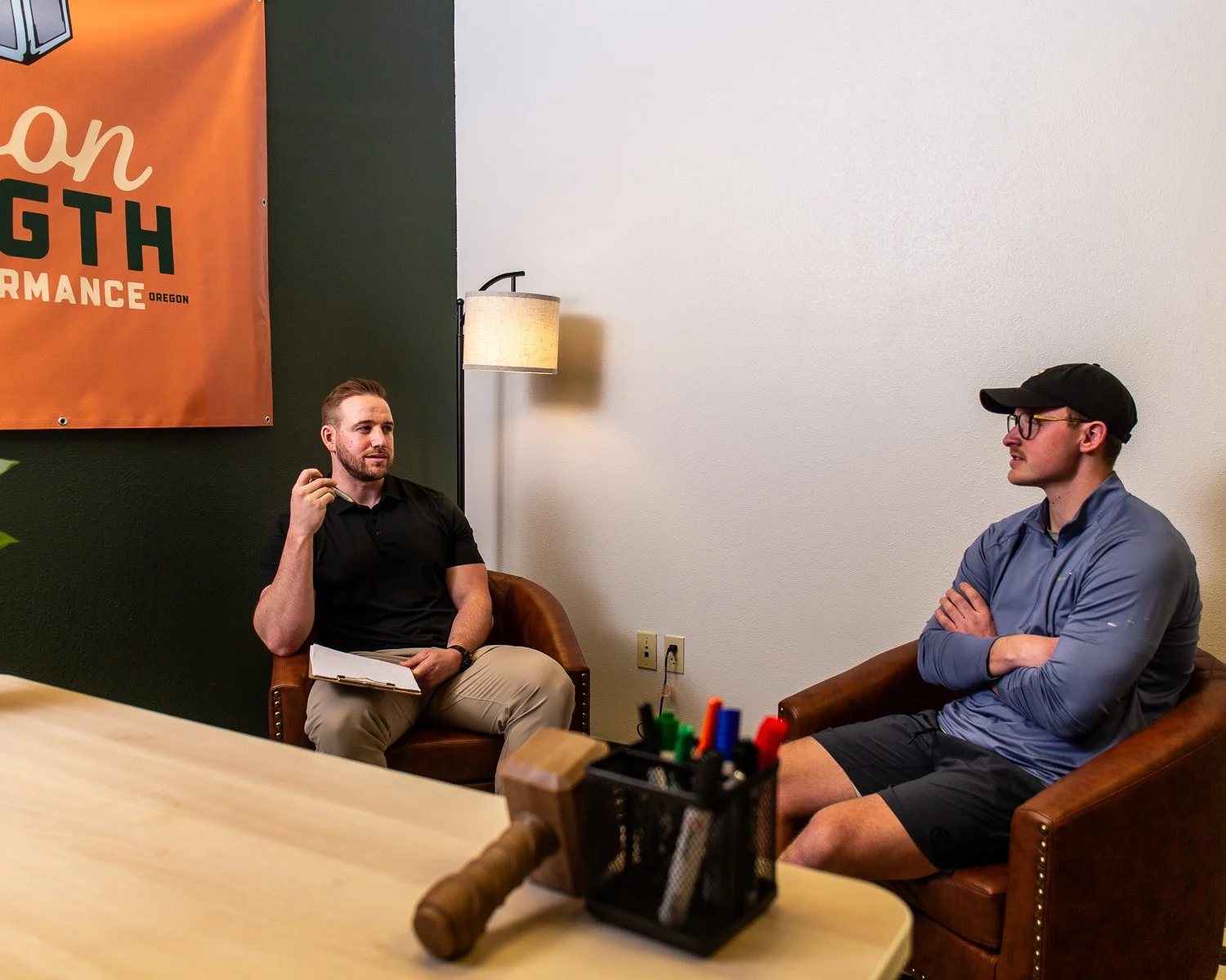 Two men having a conversation in an office setting. One is sitting on the left with papers and a pen, and the other is sitting on the right with arms crossed. A lamp, a banner, and office supplies are visible.
