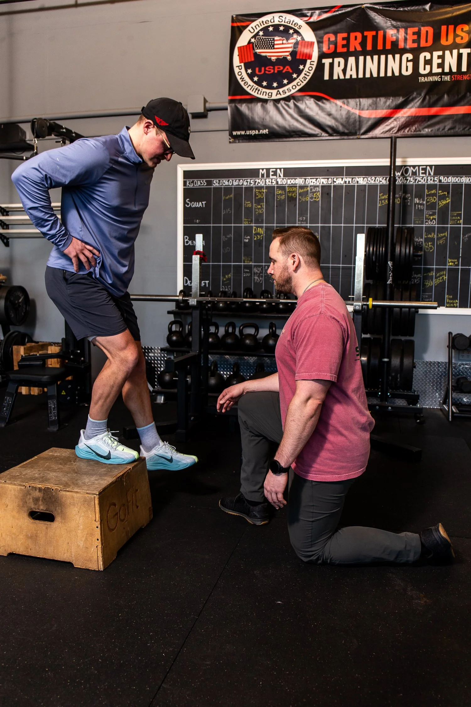 A man in a blue jacket and shorts is performing a step-up exercise on a wooden box while a trainer in a pink shirt kneels beside him, guiding him in a gym with fitness equipment and a USPA training center banner in the background.