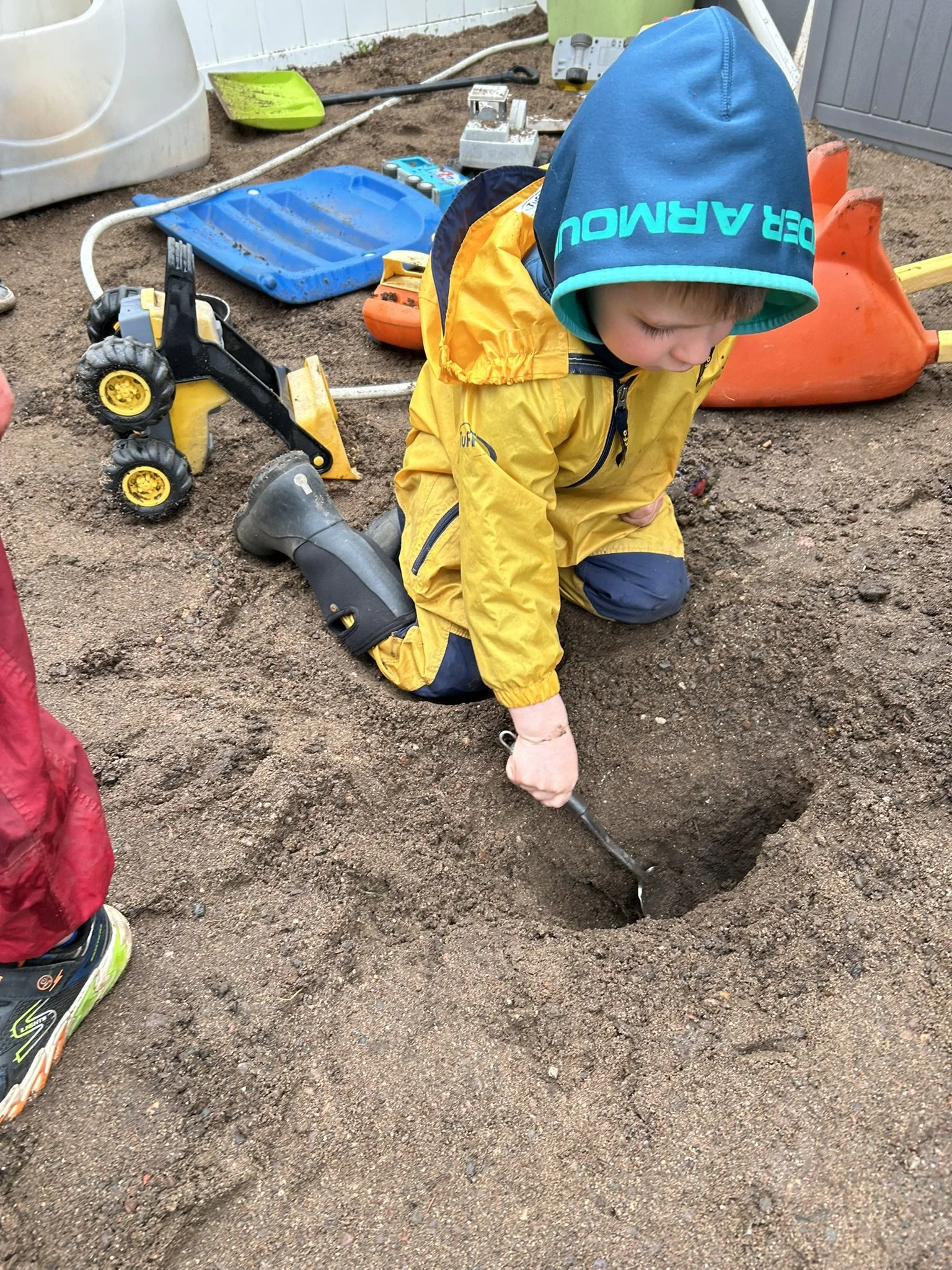 Child outside digging in the dirt with a small garden fork, surrounded by gardening tools and equipment.