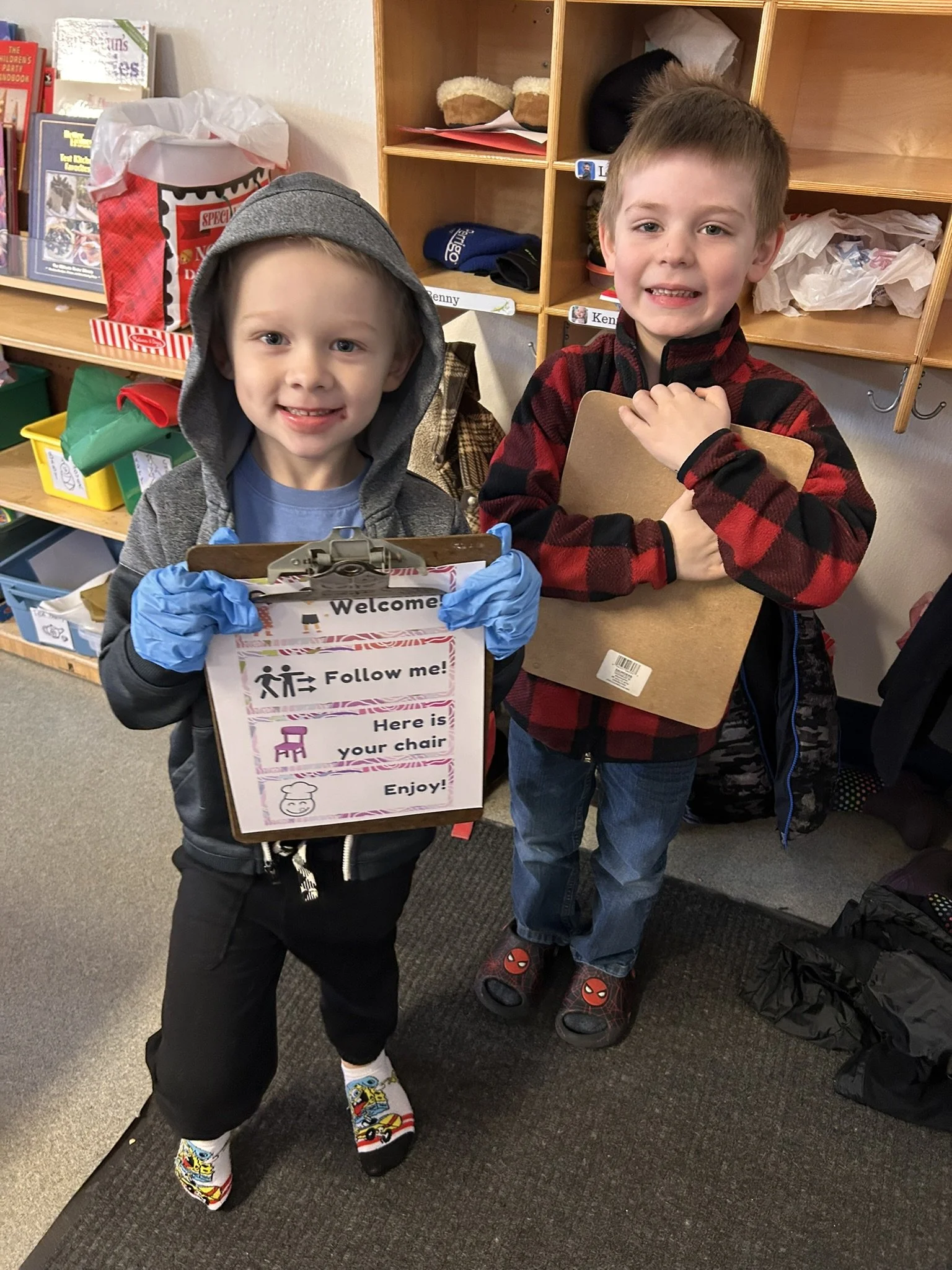 Two young boys standing in a classroom, holding clipboards. The boy on the left wears a gray hoodie with a hood over his head, black pants, and colorful socks with cars. The boy on the right wears a red and black checkered jacket, blue jeans, and slippers with Spider-Man logo. The boy on the left has a sign saying 'Welcome', 'Follow me!', 'Here is your chair', and 'Enjoy!' with illustrations, while the boy on the right holds a plain clipboard.
