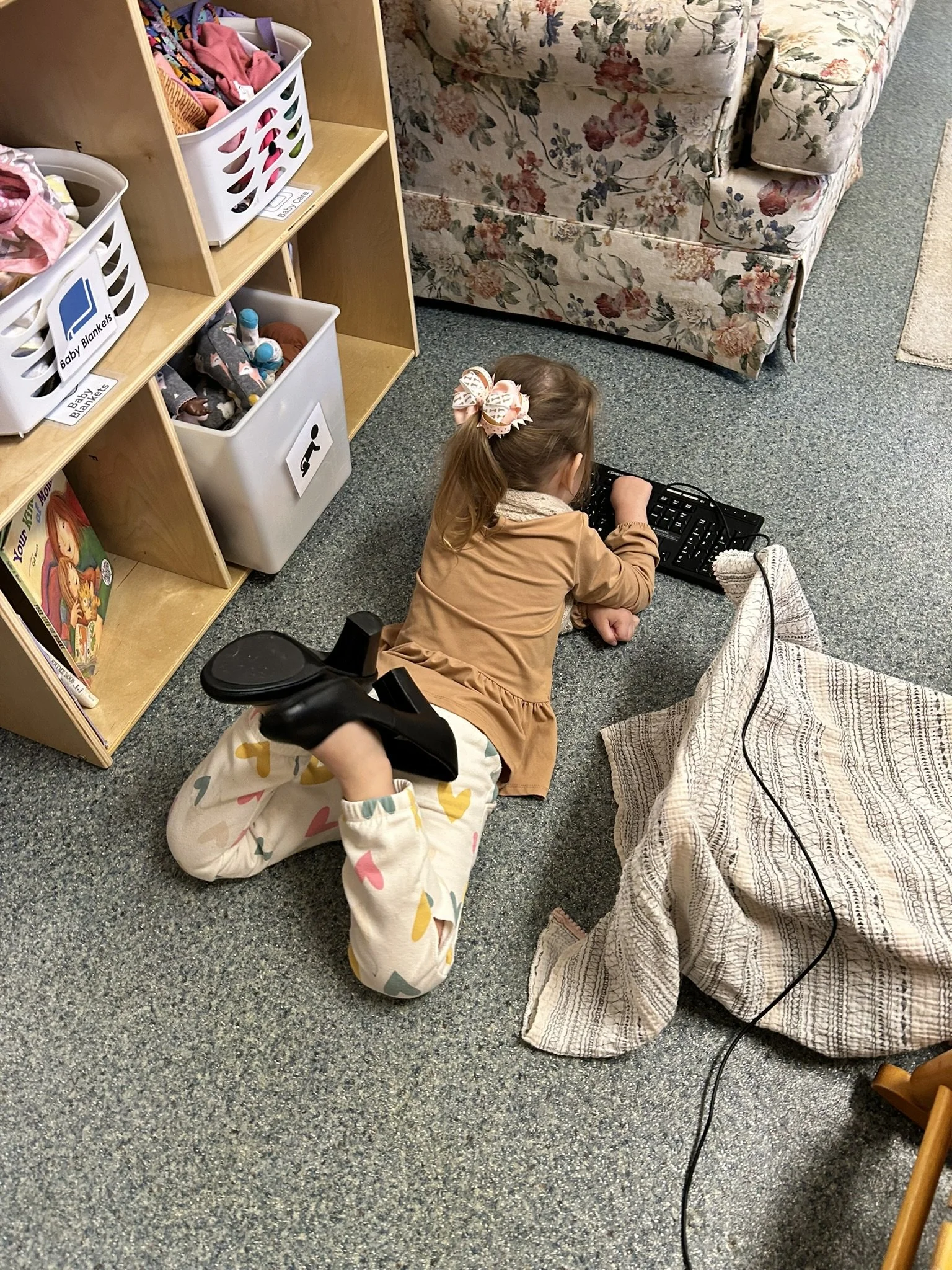 A young girl with a bow in her hair, wearing a beige top and colorful heart-patterned pants, lies on her stomach on a gray carpeted floor, playing with a small electronic keyboard.