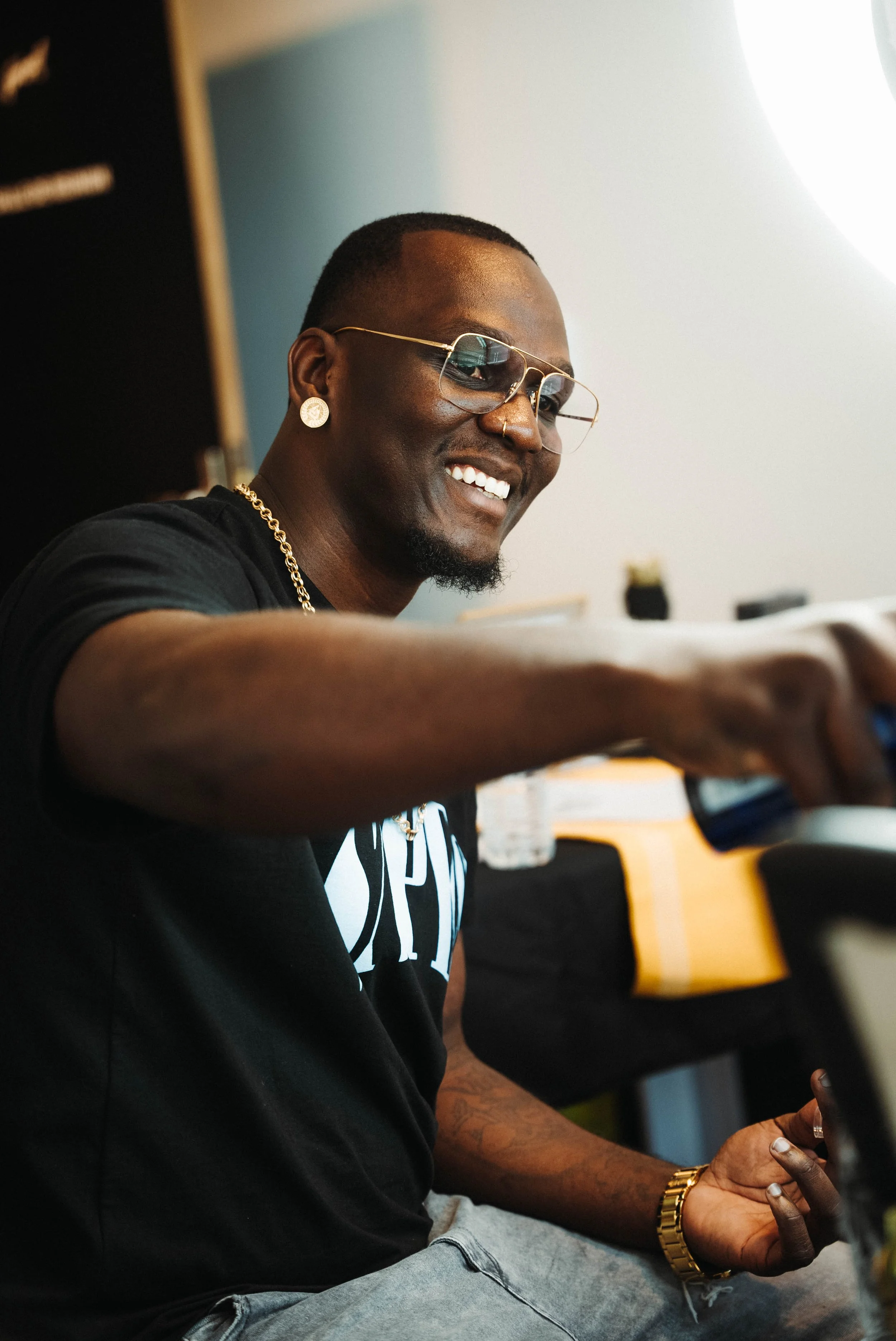 Homme souriant portant des lunettes de soleil, une chaîne en or, une boucle d'oreille et un bracelet en or, assis à une table