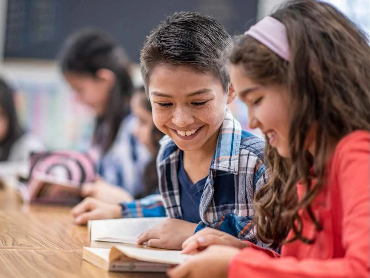 Two smiling elementary students reading a book together at a wooden desk in a bright classroom setting, with other classmates working in the blurred background.