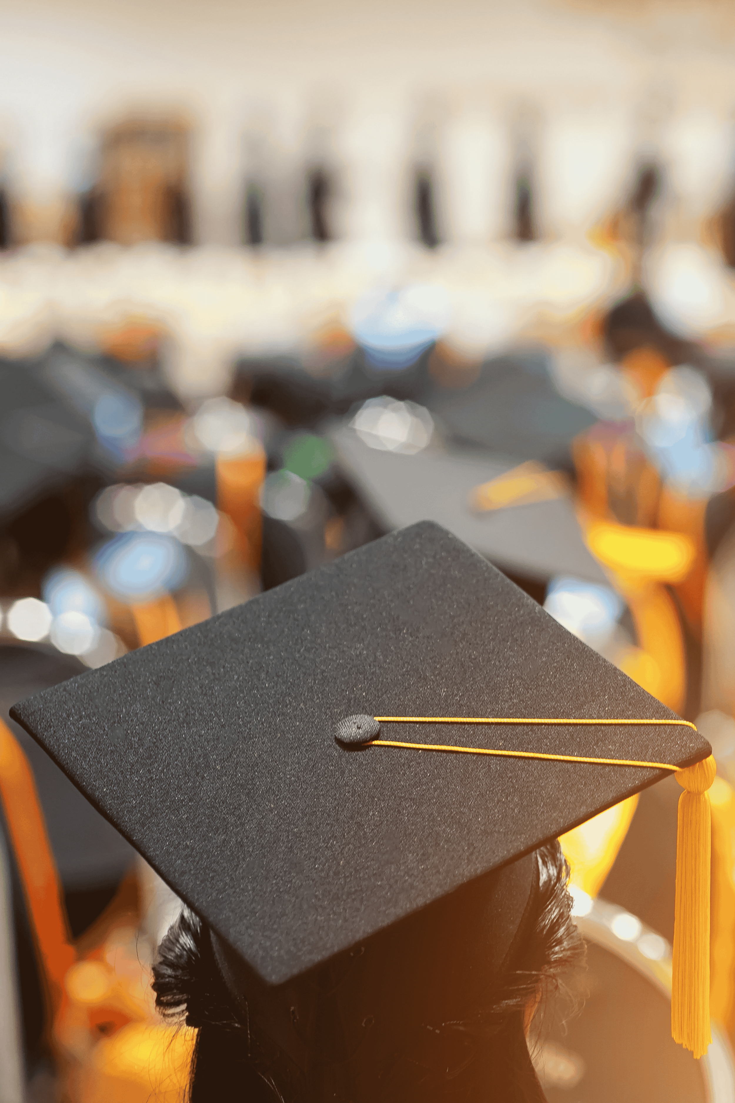 Graduate student wearing a black mortarboard cap with a golden tassel looking toward a group of graduates at a ceremony.