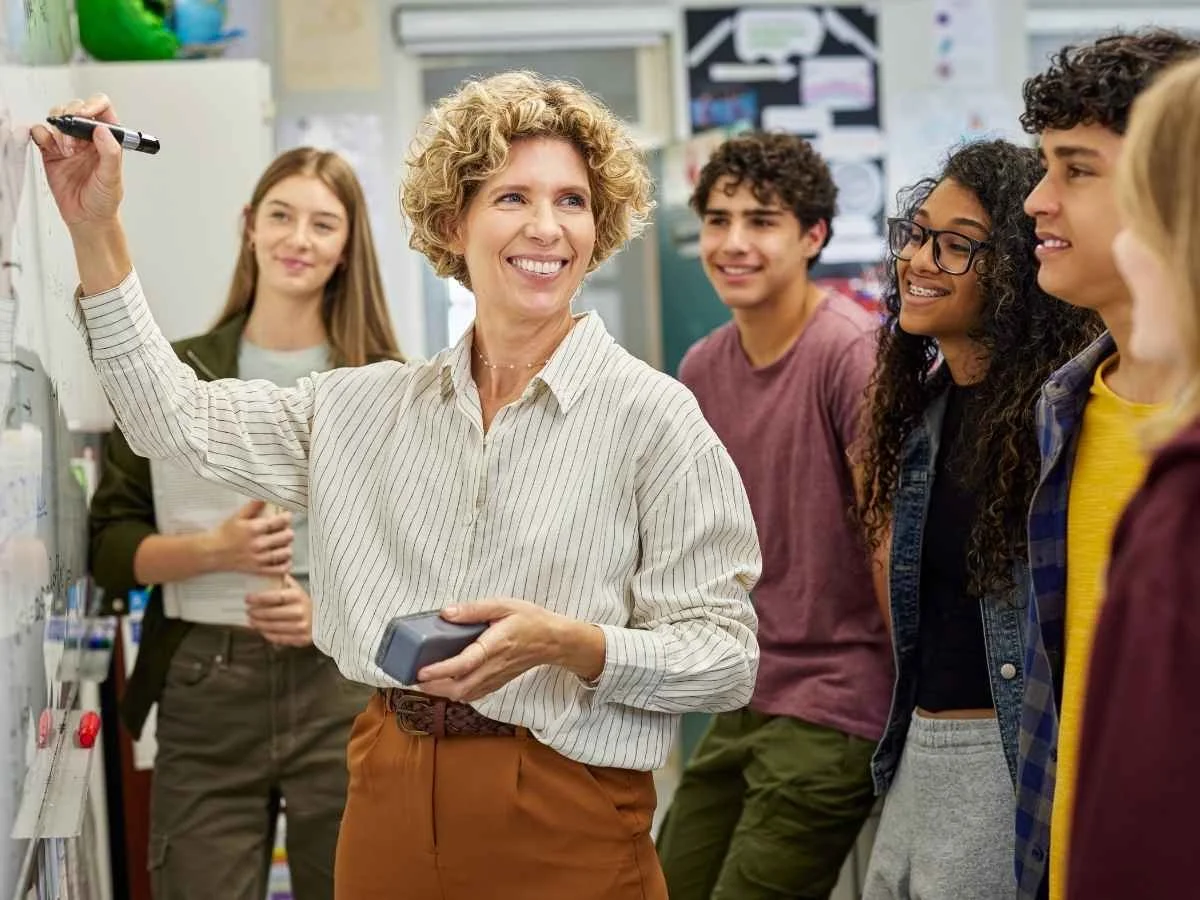 A smiling, curly-haired teacher writes on a whiteboard while a diverse group of teenage students looks on with engagement in a bright classroom.