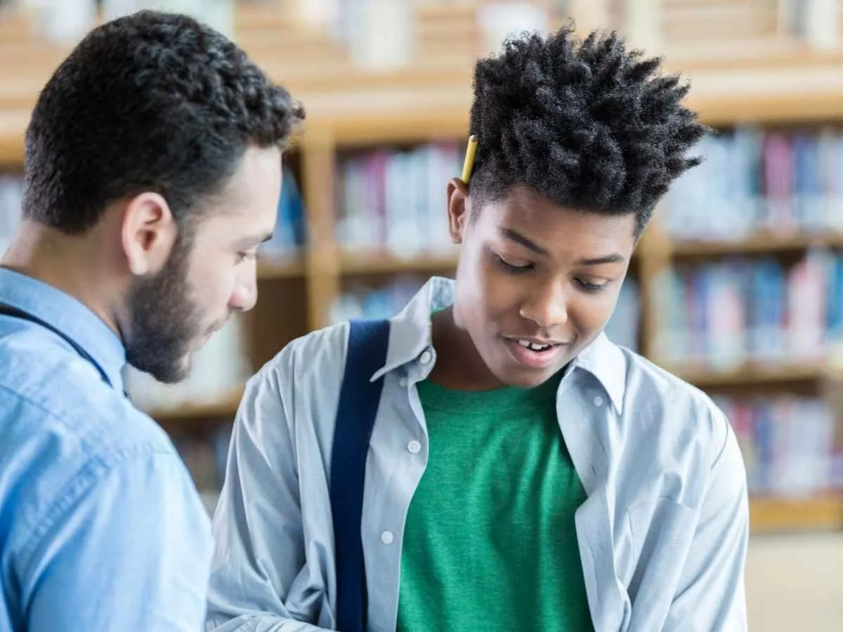 A male educator and a young male student standing together in a bright setting, both looking down and smiling at an open textbook held by the student.