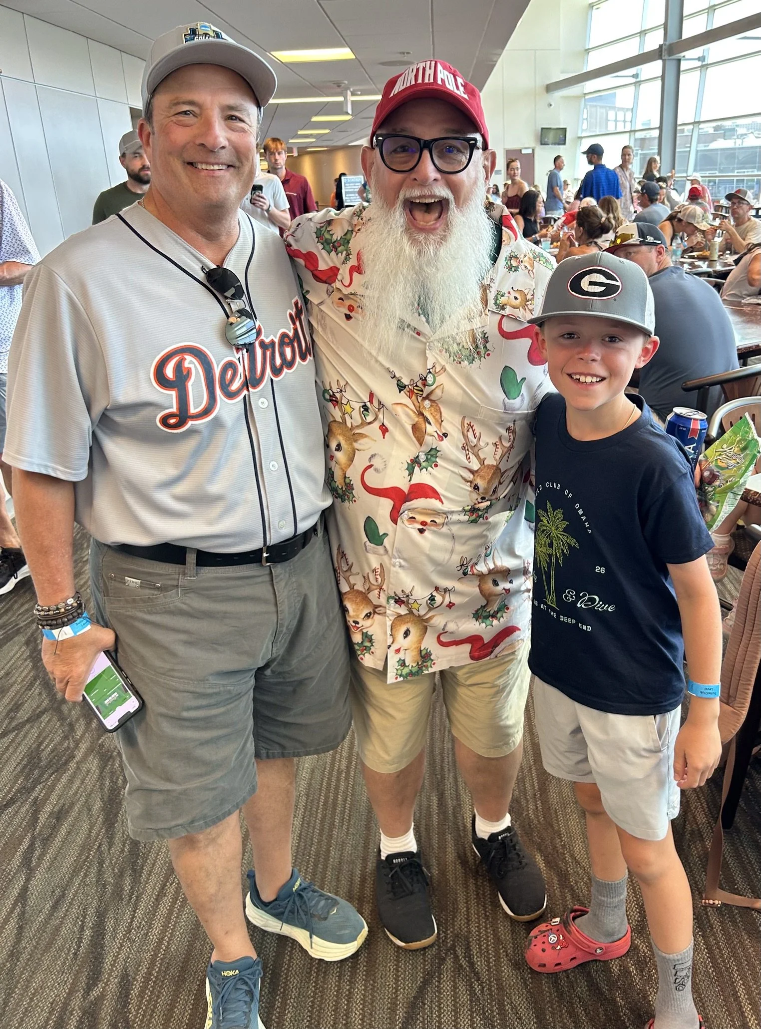 Three smiling males standing together at an indoor event with many seated and standing people in the background. One wearing a Detroit Tigers baseball jersey, another in a Christmas-themed shirt and red North Pole cap, and a young boy with a Georgia 