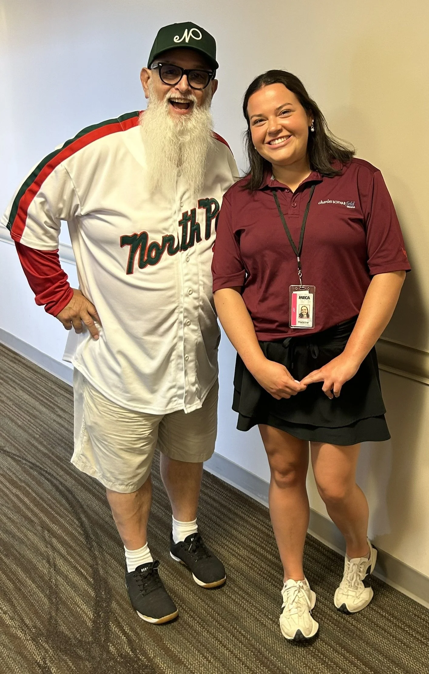 A man wearing a North Platte baseball jersey, khaki shorts, black sneakers, and a cap next to a woman in a maroon shirt with a name badge, black skirt, and white sneakers. They are smiling and standing indoors against a plain wall.
