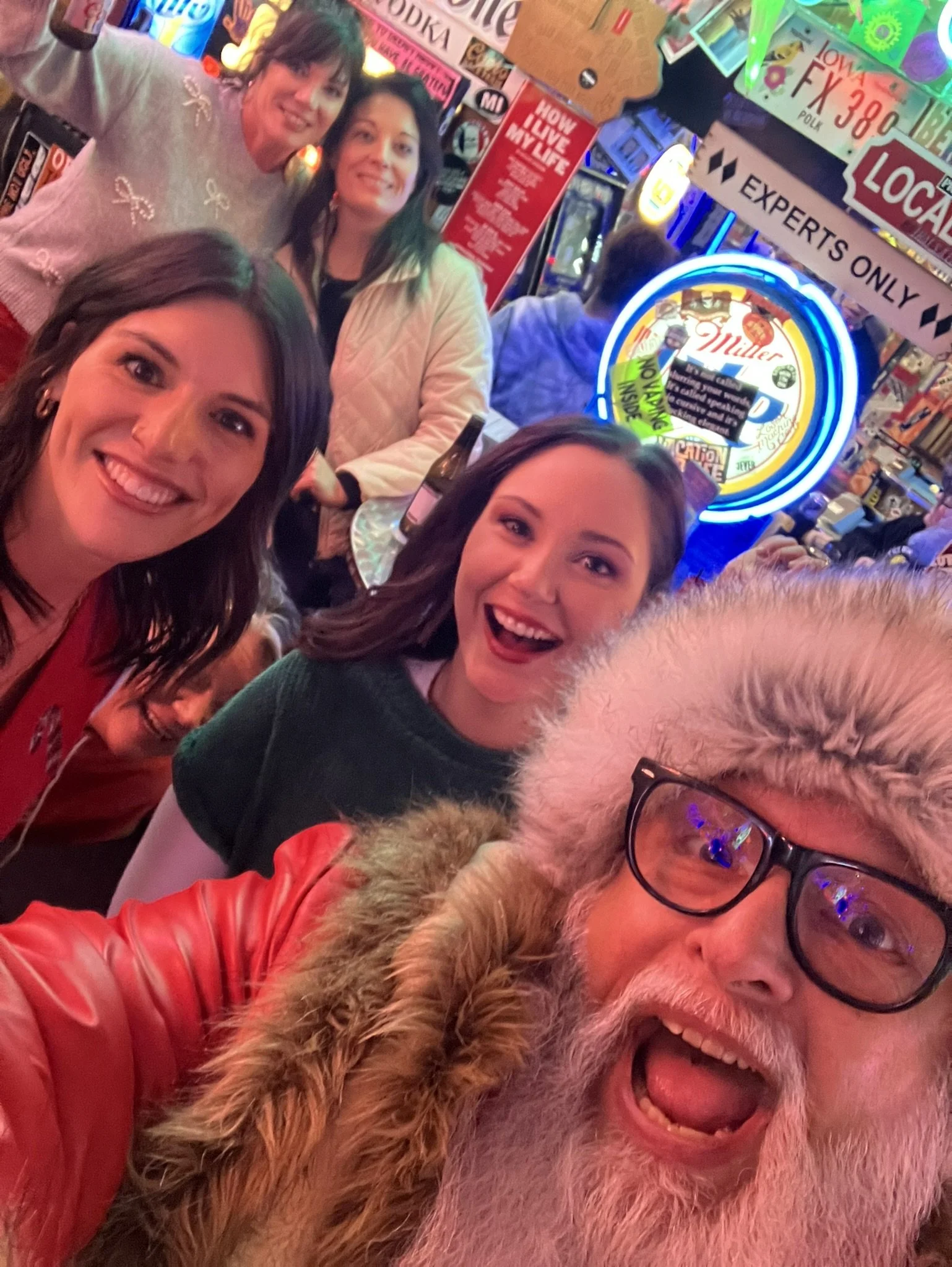 A group of people in a lively bar or pub, smiling and taking a selfie. The background has colorful neon signs and advertisements, including a large Miller Lite sign. The atmosphere appears fun and festive.