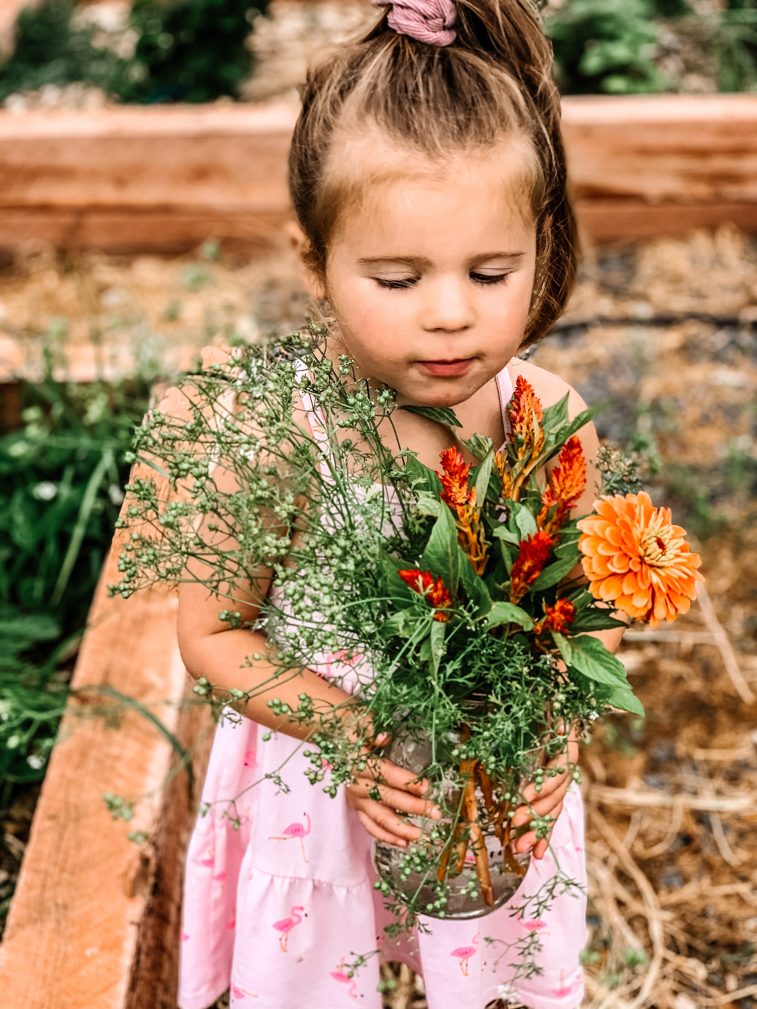 A young girl with brown hair in a pink dress holding a bouquet of colorful flowers outdoors.