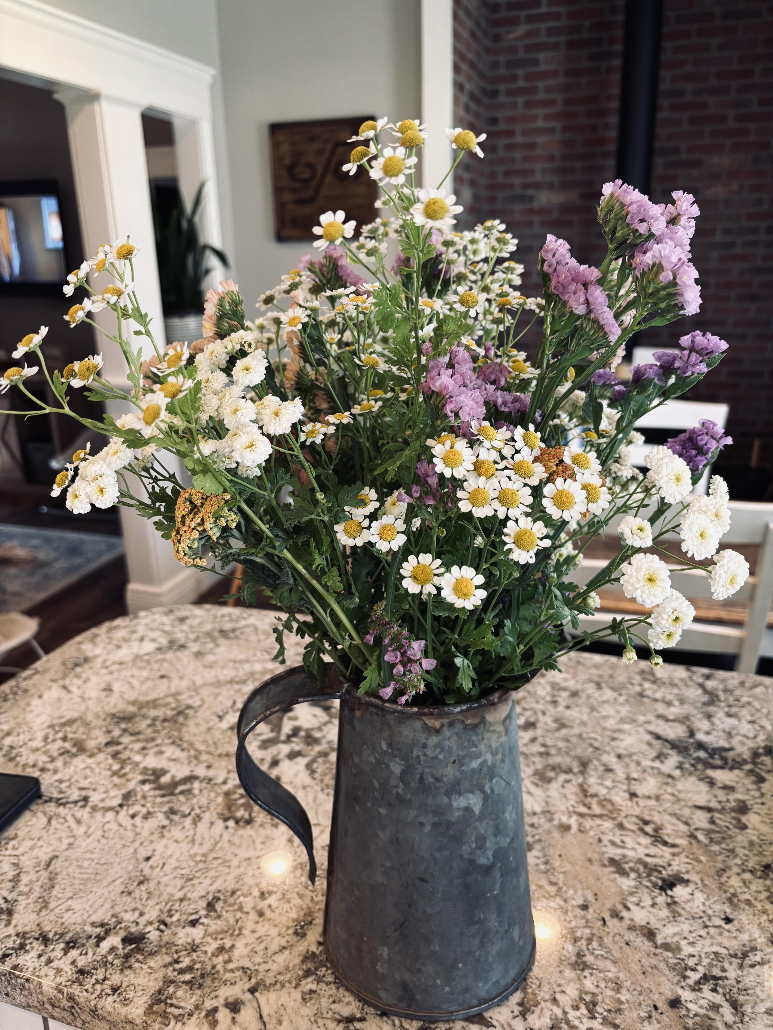 A rustic metal pitcher filled with a bouquet of white, yellow, and purple wildflowers on a kitchen countertop.