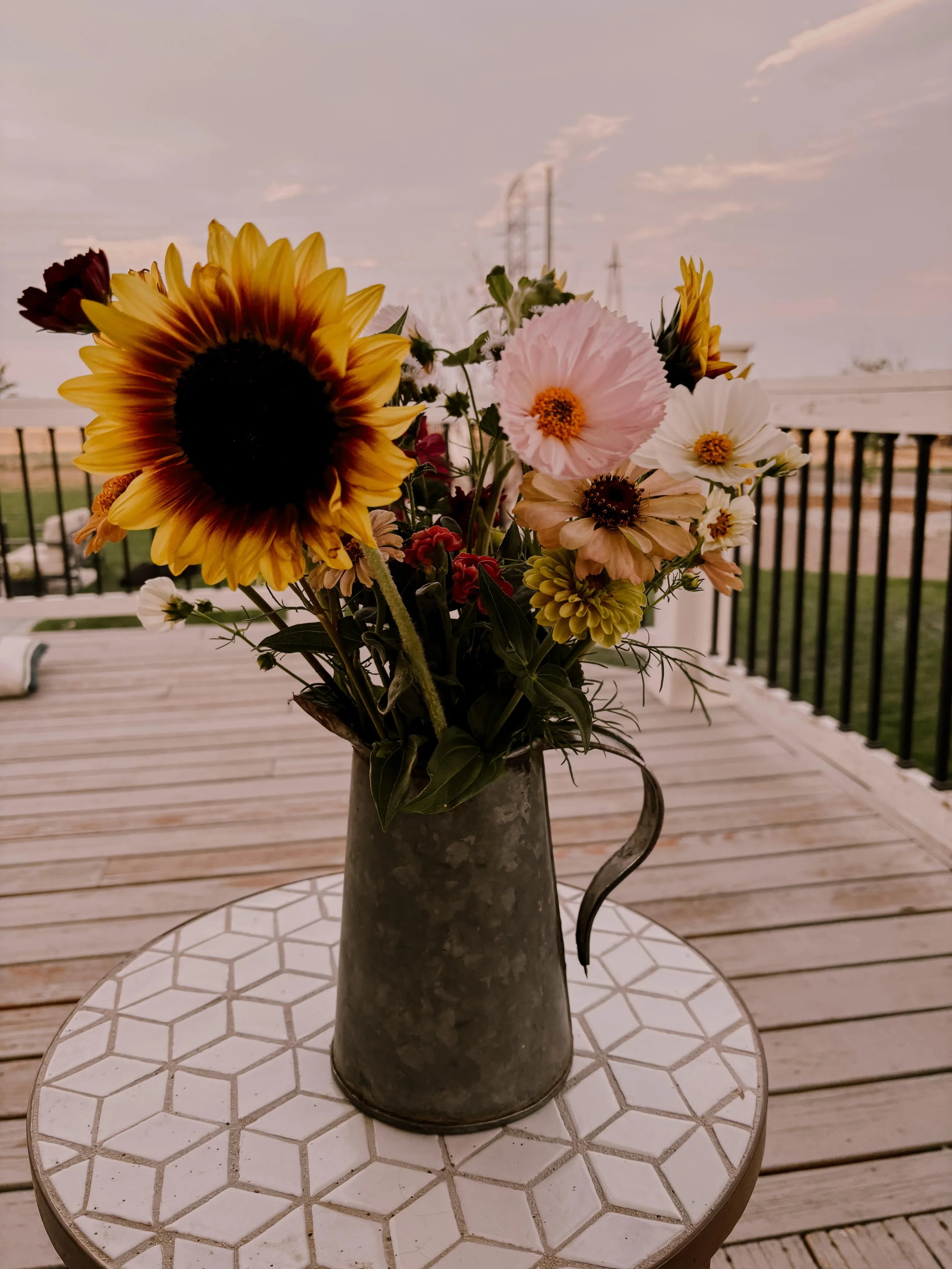 A metal pitcher vase containing a bouquet of colorful flowers, including sunflowers with large yellow petals and black centers, pink, white, and beige flowers, placed on a small white table with a hexagon pattern. The background shows a wooden deck with black railing and a partly cloudy sky.