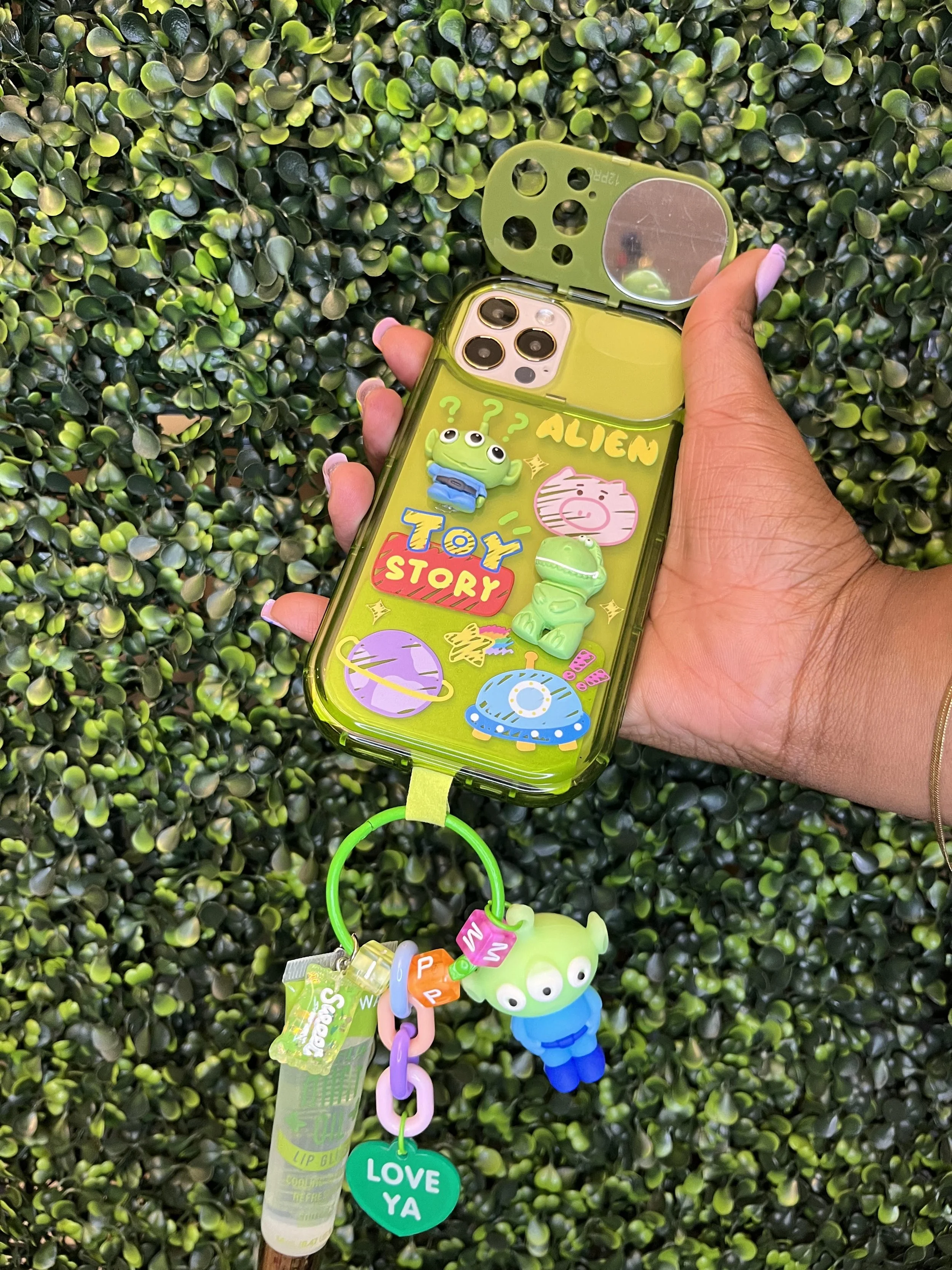 A hand holding a green phone case decorated with Toy Story stickers, including aliens, a pig, and a spaceship, attached to a keychain with a green alien figure and a green heart-shaped tag with "LOVE YA" written on it, against a background of dense g