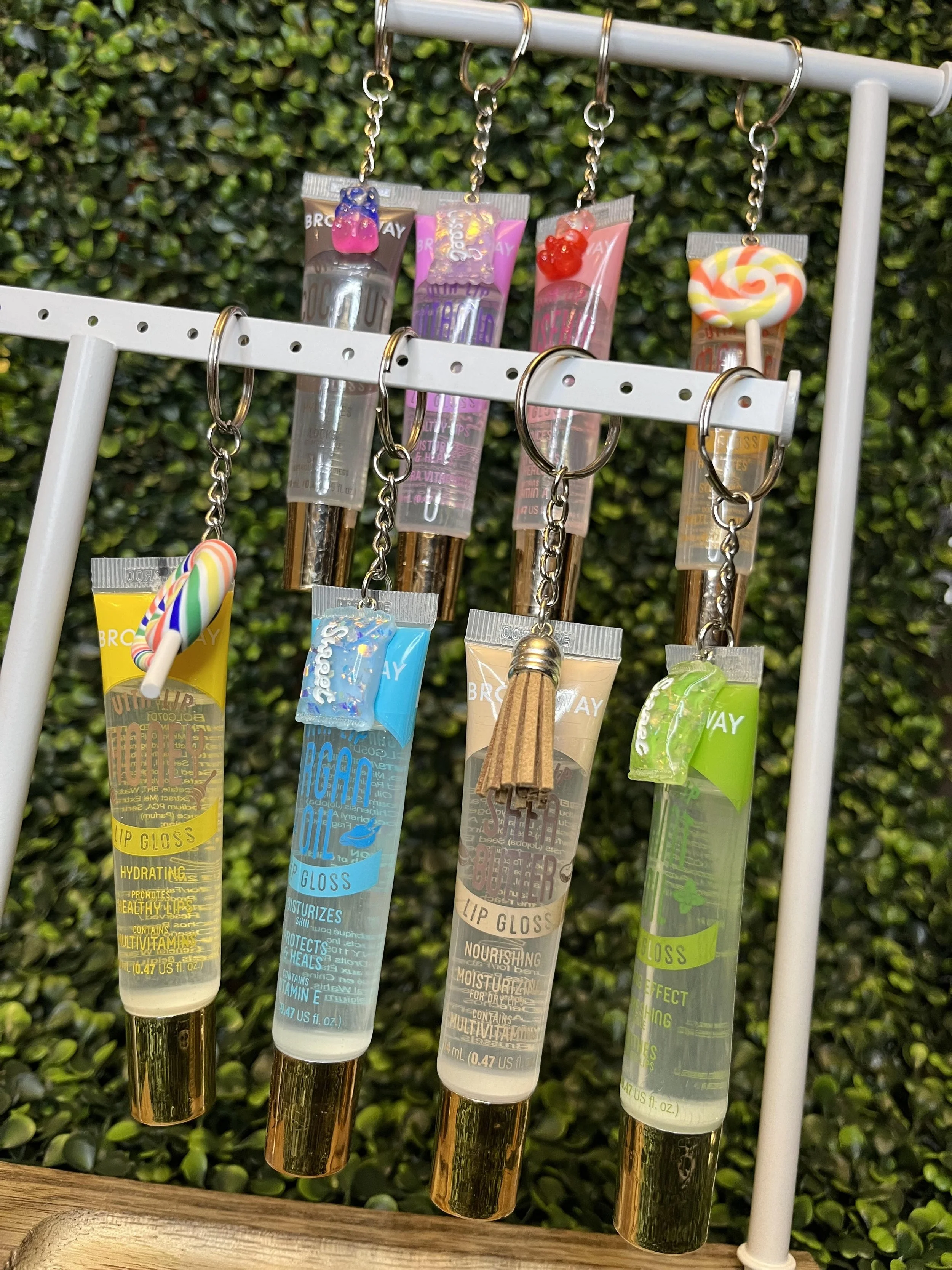 Set of lip gloss keychains with small toy shapes on tops, hanging from a white display rack, with a green leafy background.
