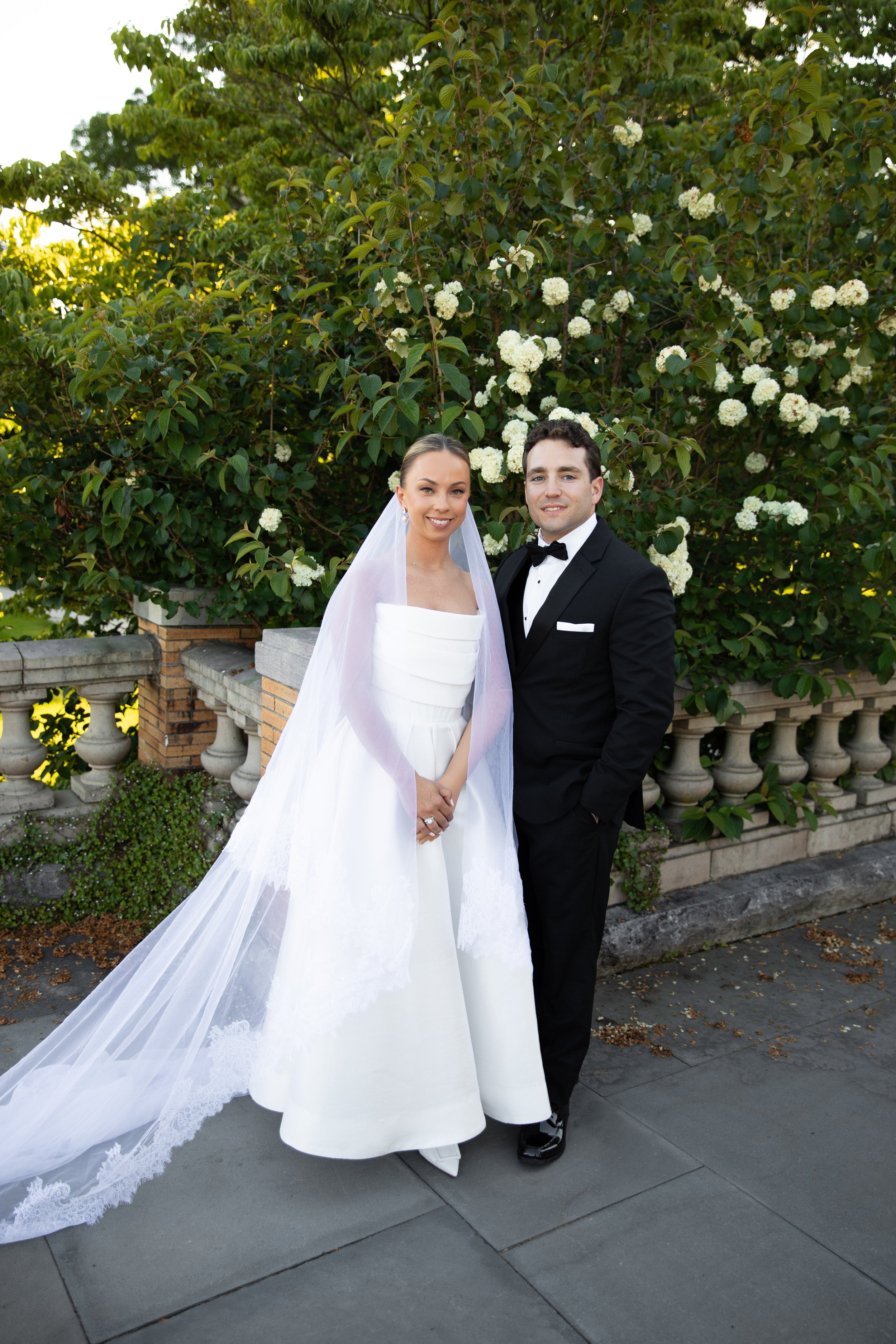 A bride and groom standing outdoors in front of green bushes with white flowers, dressed in wedding attire, smiling at the camera.