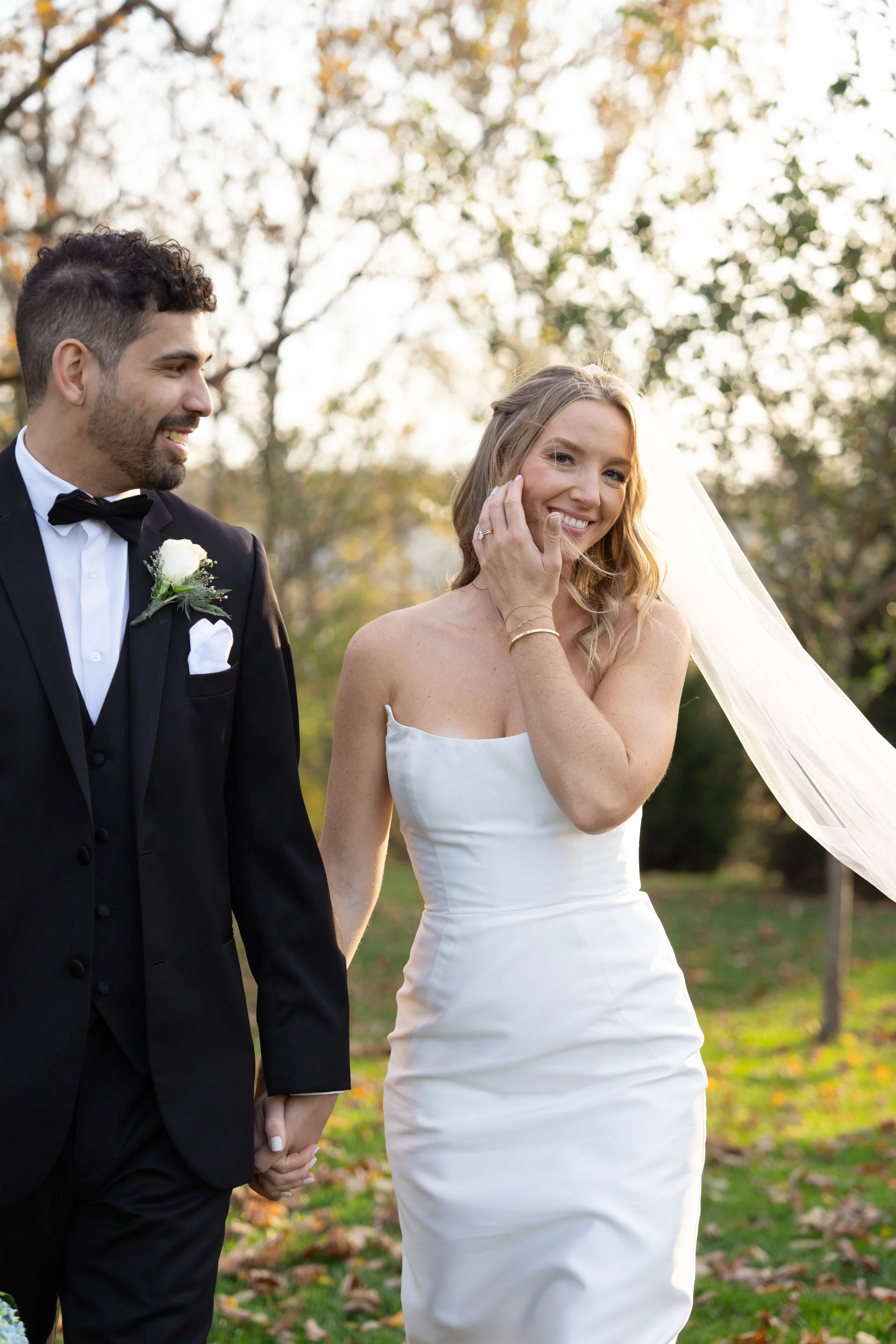 A bride and groom holding hands outdoors during a wedding, with the bride smiling and touching her face, dressed in a white wedding gown, and the groom in a black tuxedo, both surrounded by autumn trees.
