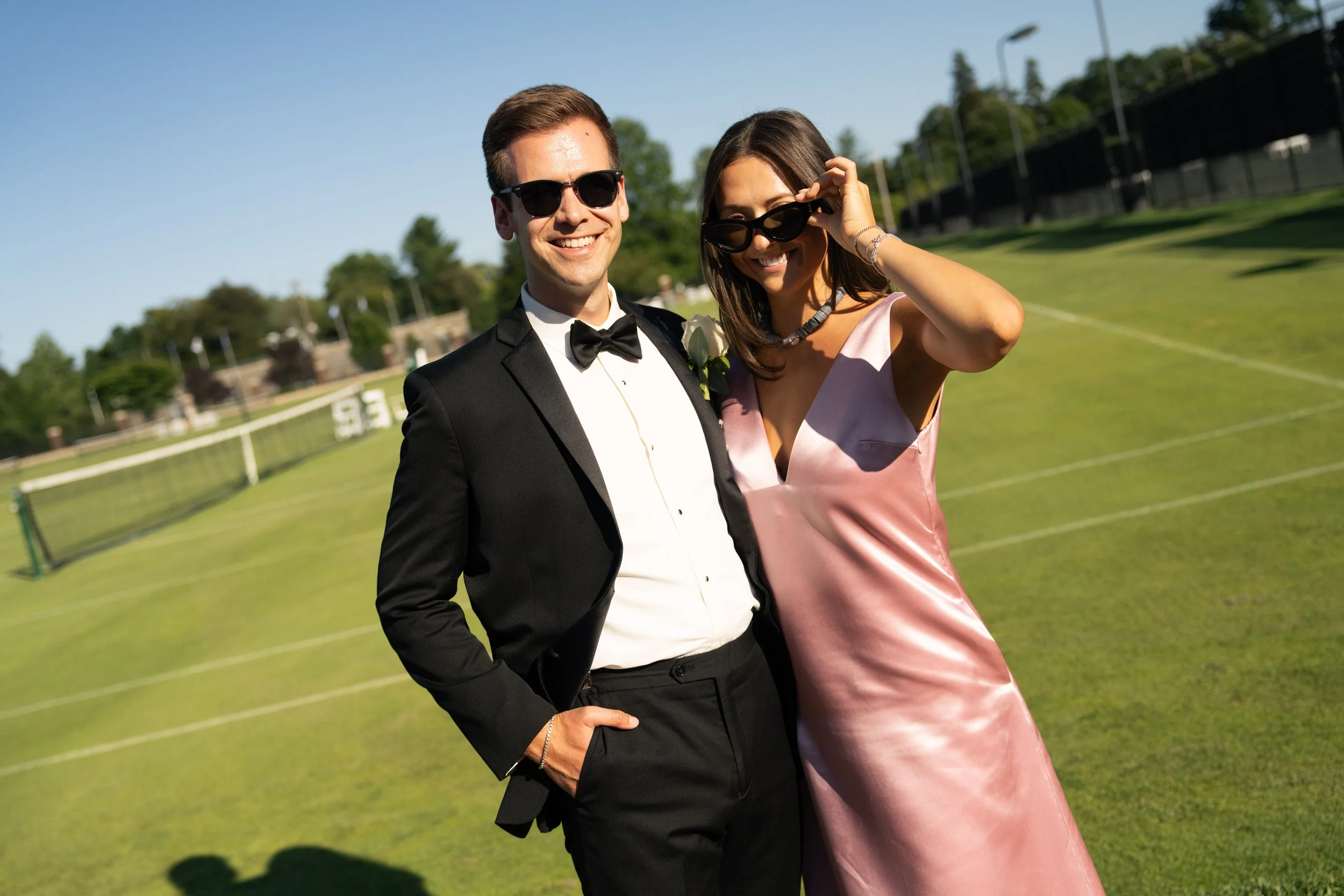 A couple dressed in formal attire, smiling and wearing sunglasses, posing outdoors on a sunny day with a tennis court in the background.