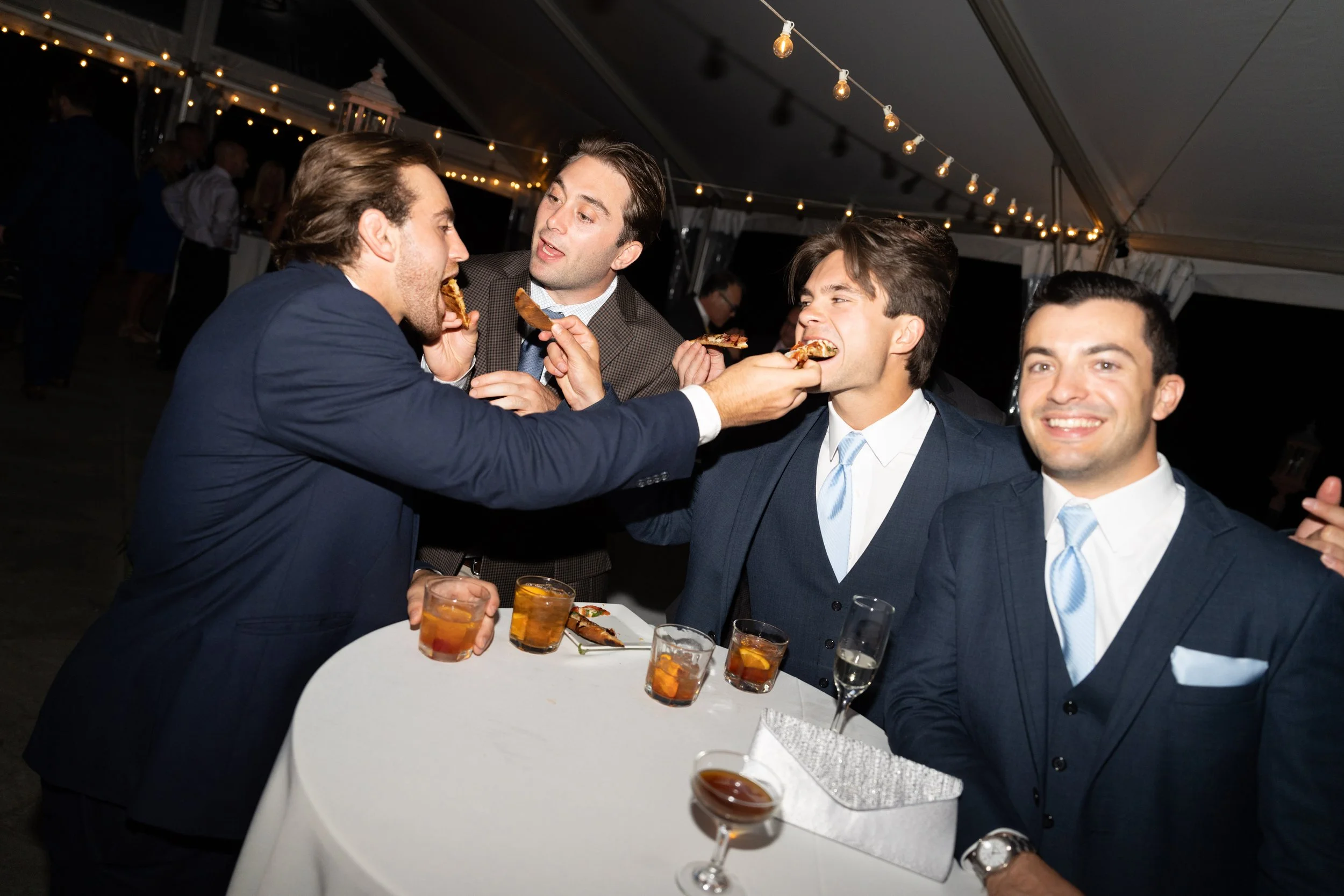 Four men in suits at a social event, some feeding each other slices of pizza, with drinks on the table and string lights overhead.