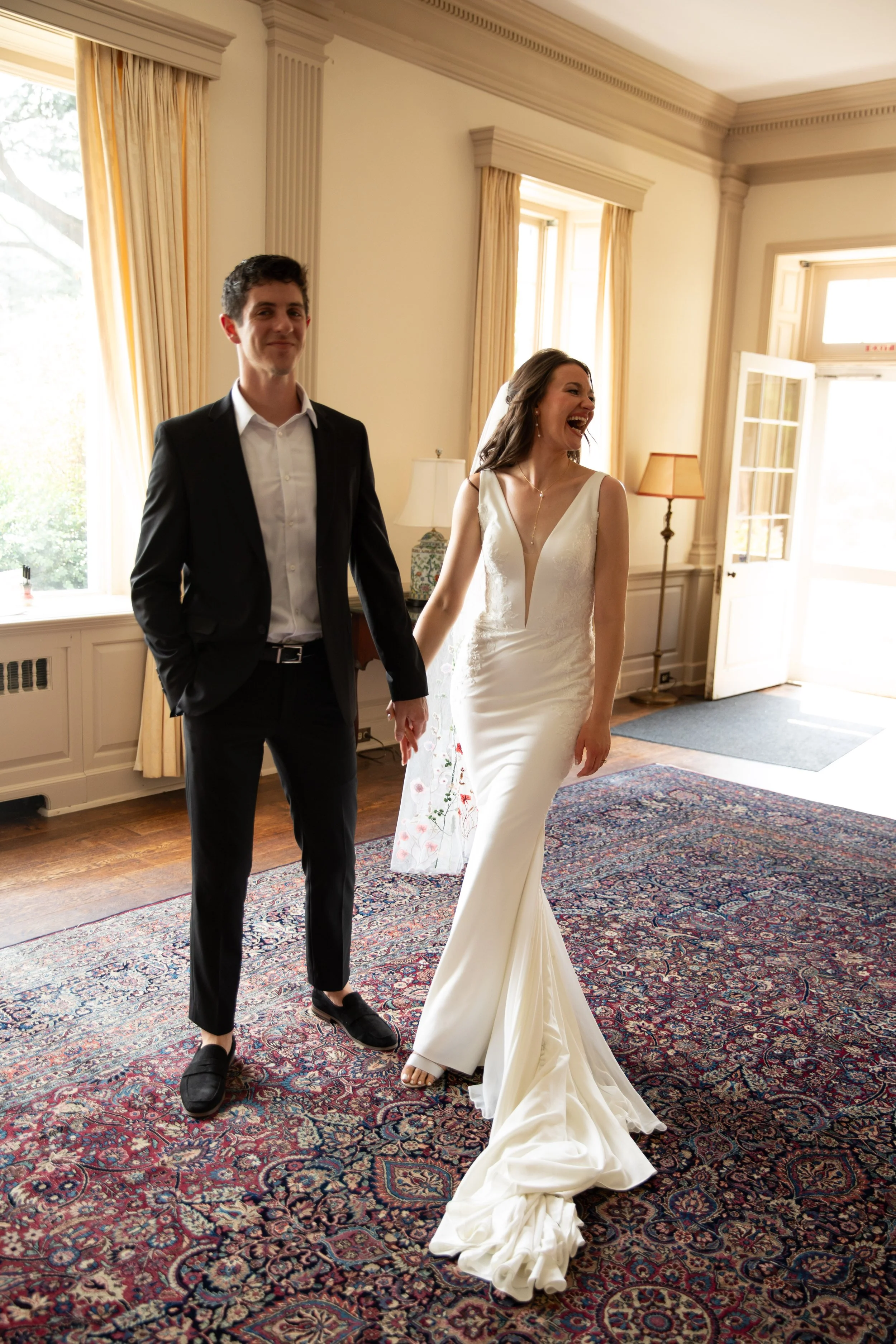 A bride and groom holding hands in a warmly lit room with large windows and patterned carpet, celebrating their wedding day.