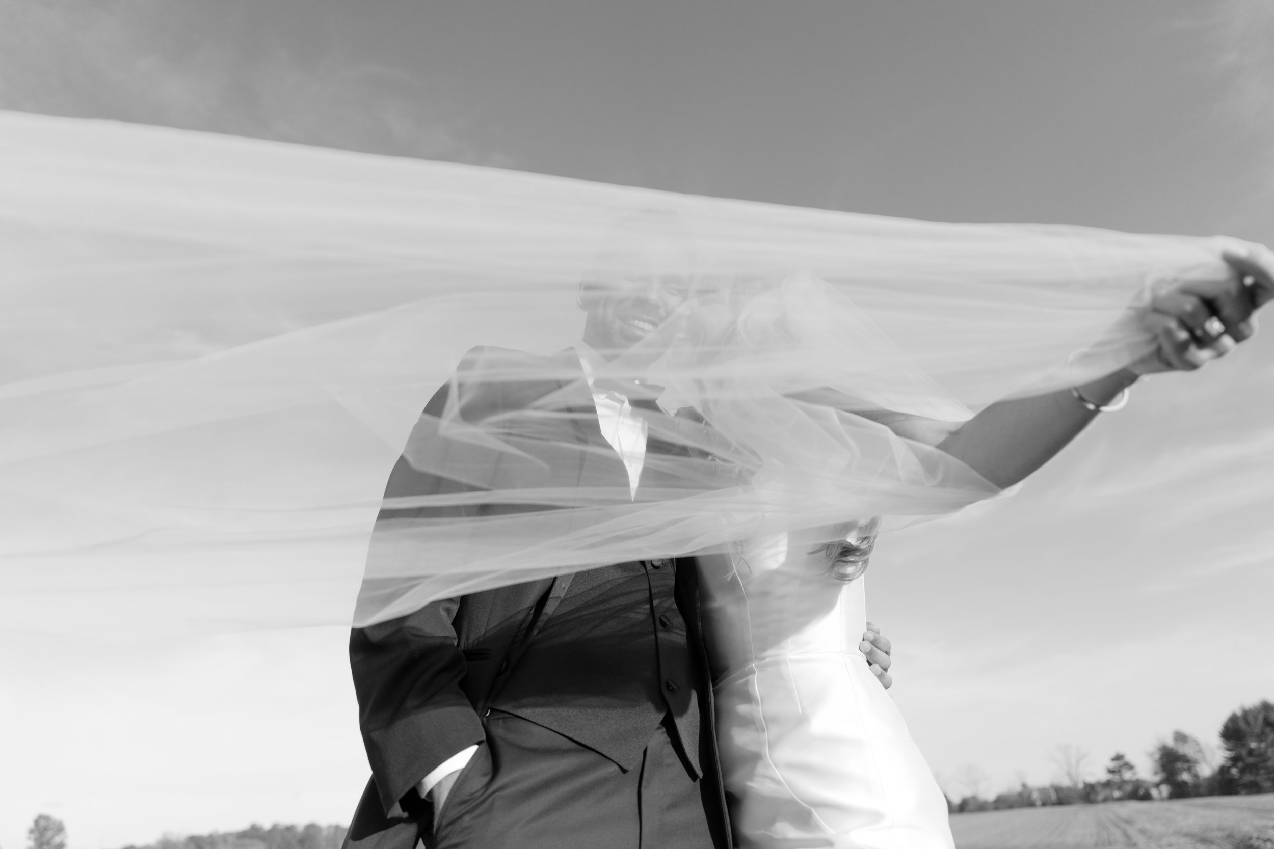 A smiling bride and groom in wedding attire, standing outdoors in an open field, with the bride's veil covering part of the groom's face in a black and white photograph.
