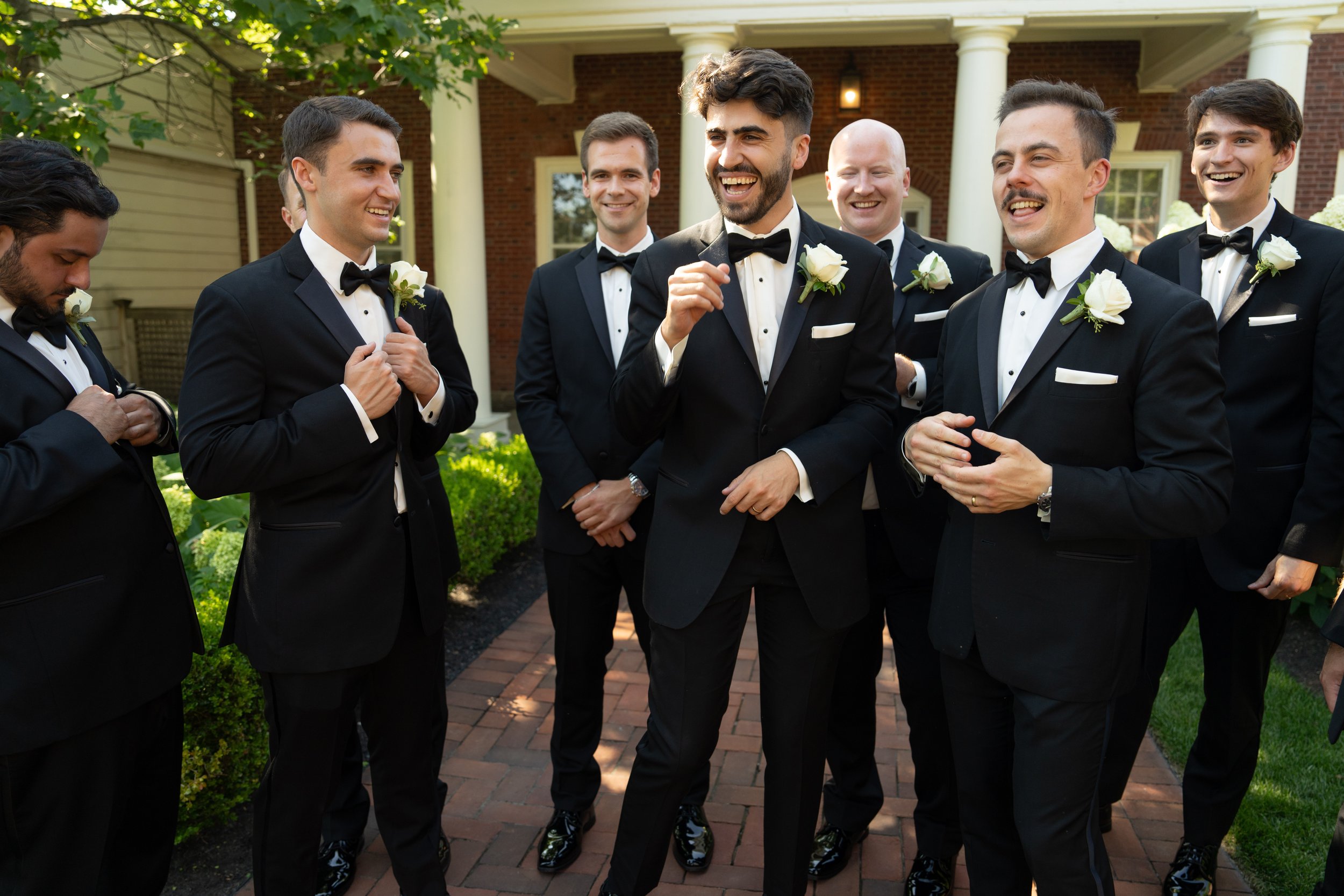Group of men in tuxedos with white roses, standing outside, smiling and talking.