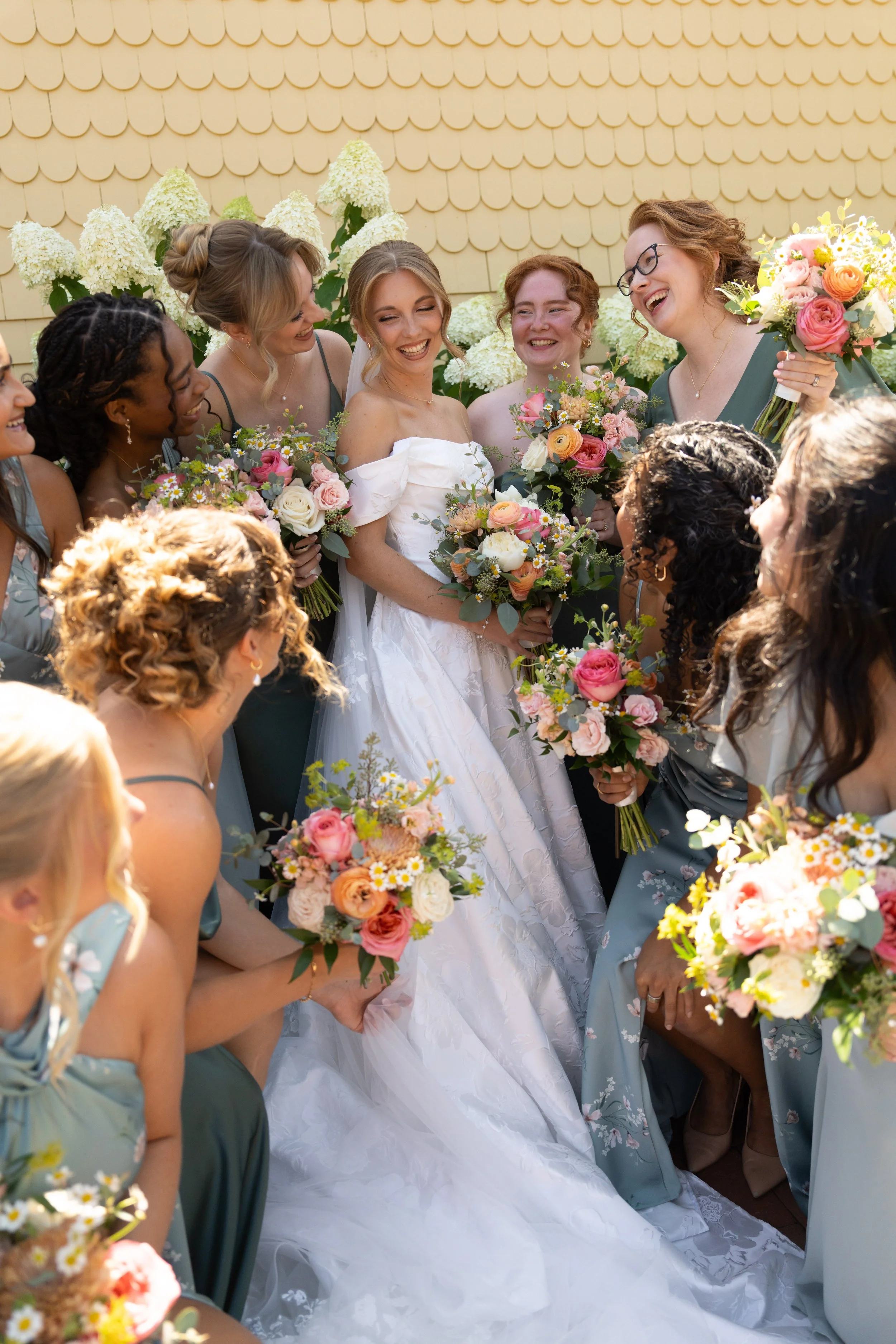 A bride in a white dress surrounded by bridesmaids in teal dresses, all holding colorful bouquets, smiling and celebrating outdoors.