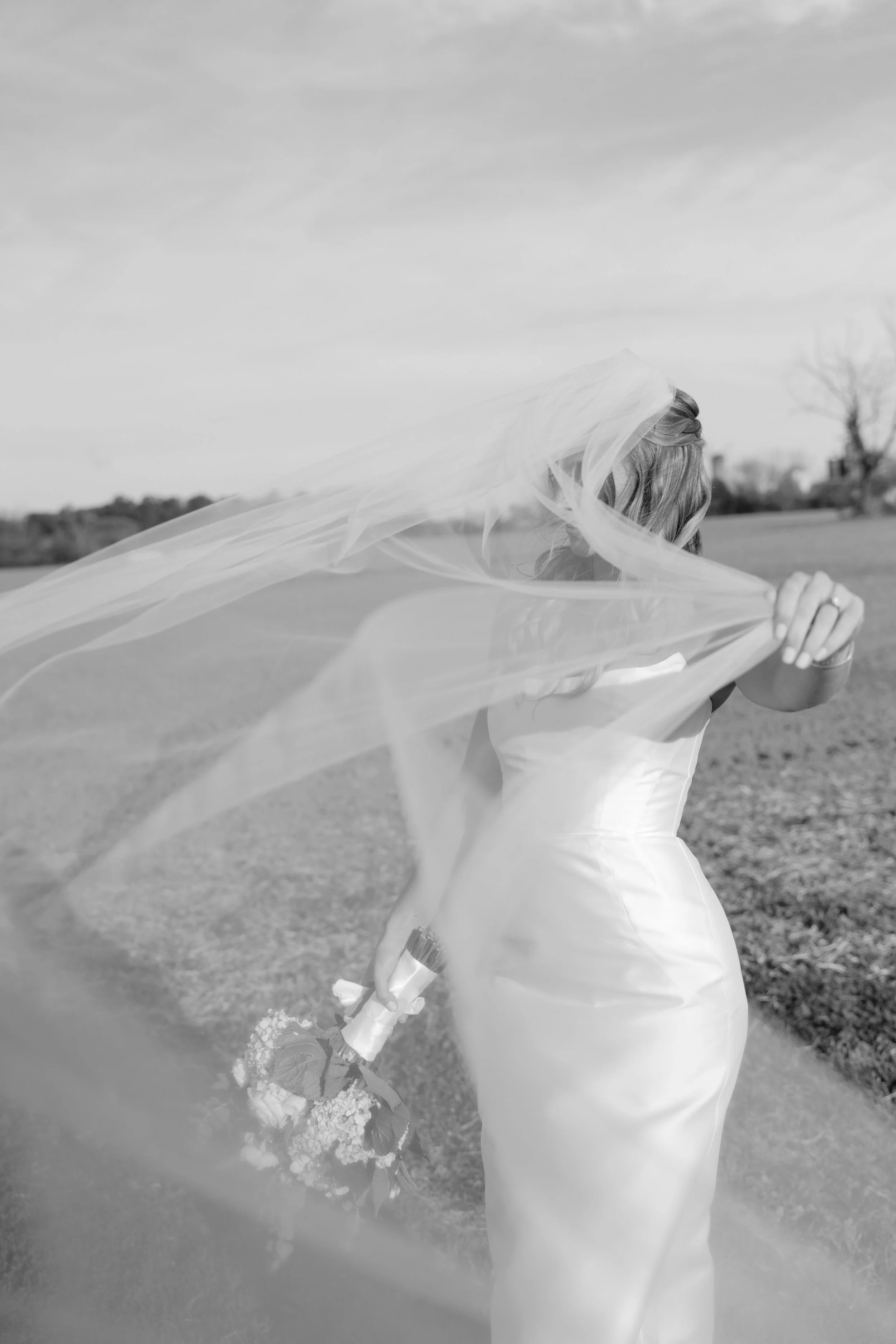 Black and white photo of a woman in a wedding dress holding a bouquet with her veil blowing in the wind, standing outdoors in a field with trees in the background.