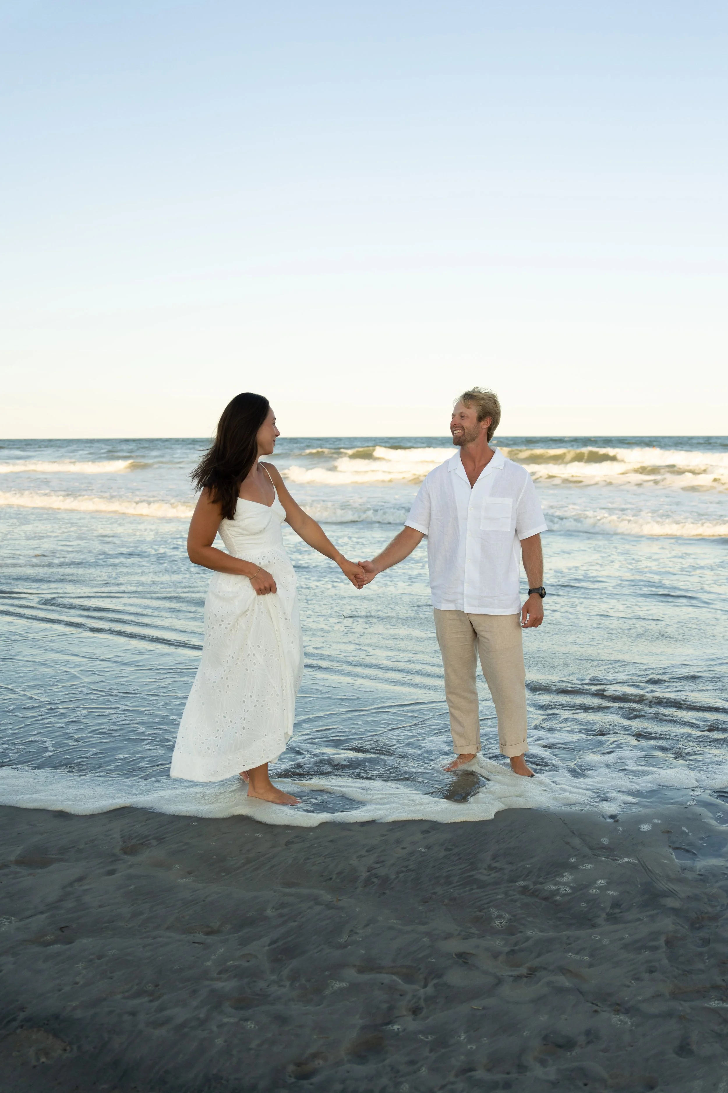 A couple holding hands in the ocean at the beach, smiling at each other, dressed in light-colored clothing with waves and the horizon in the background.