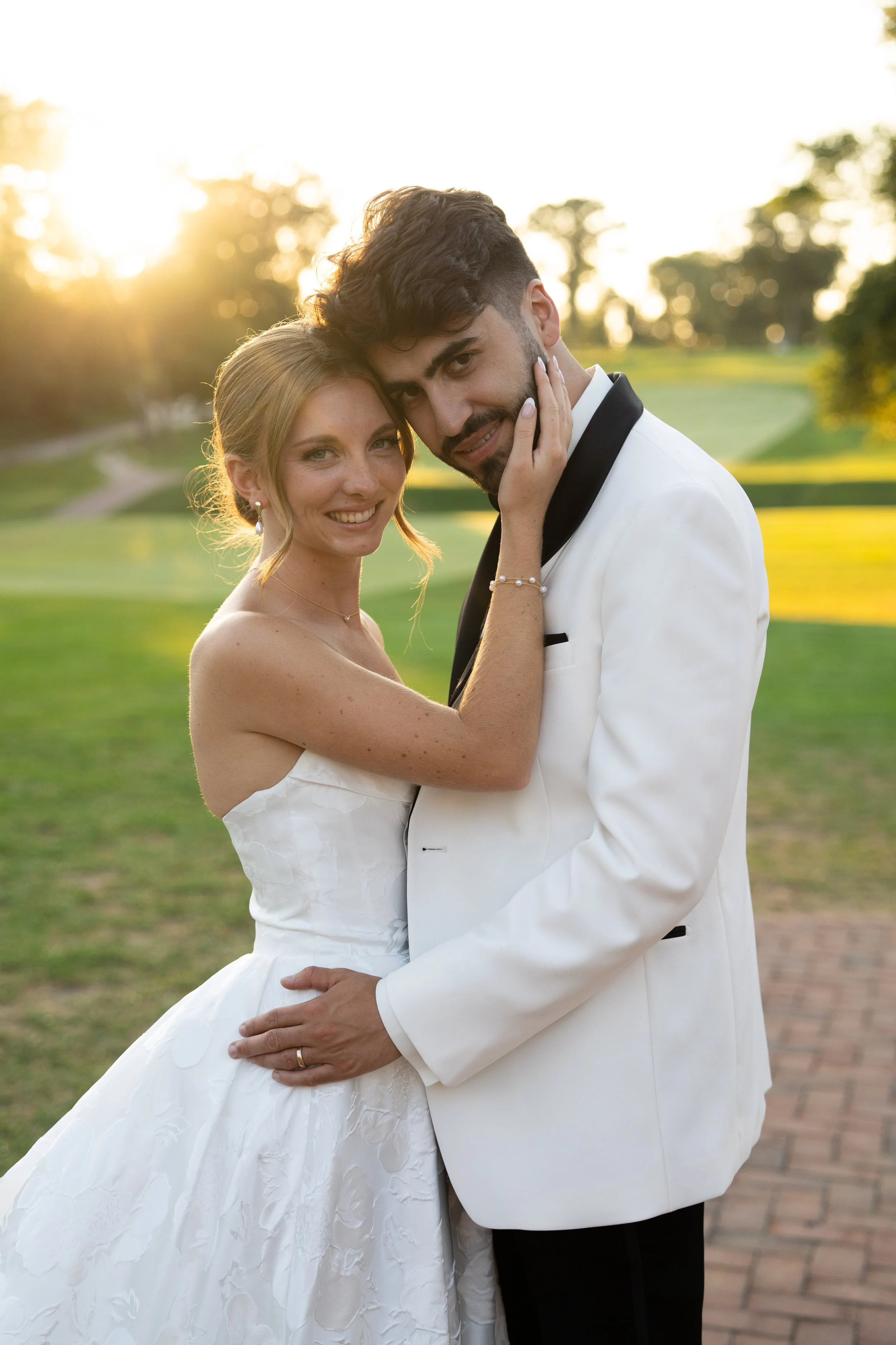 A newlywed couple in wedding attire, embracing outdoors during sunset, with green grass and trees in the background.
