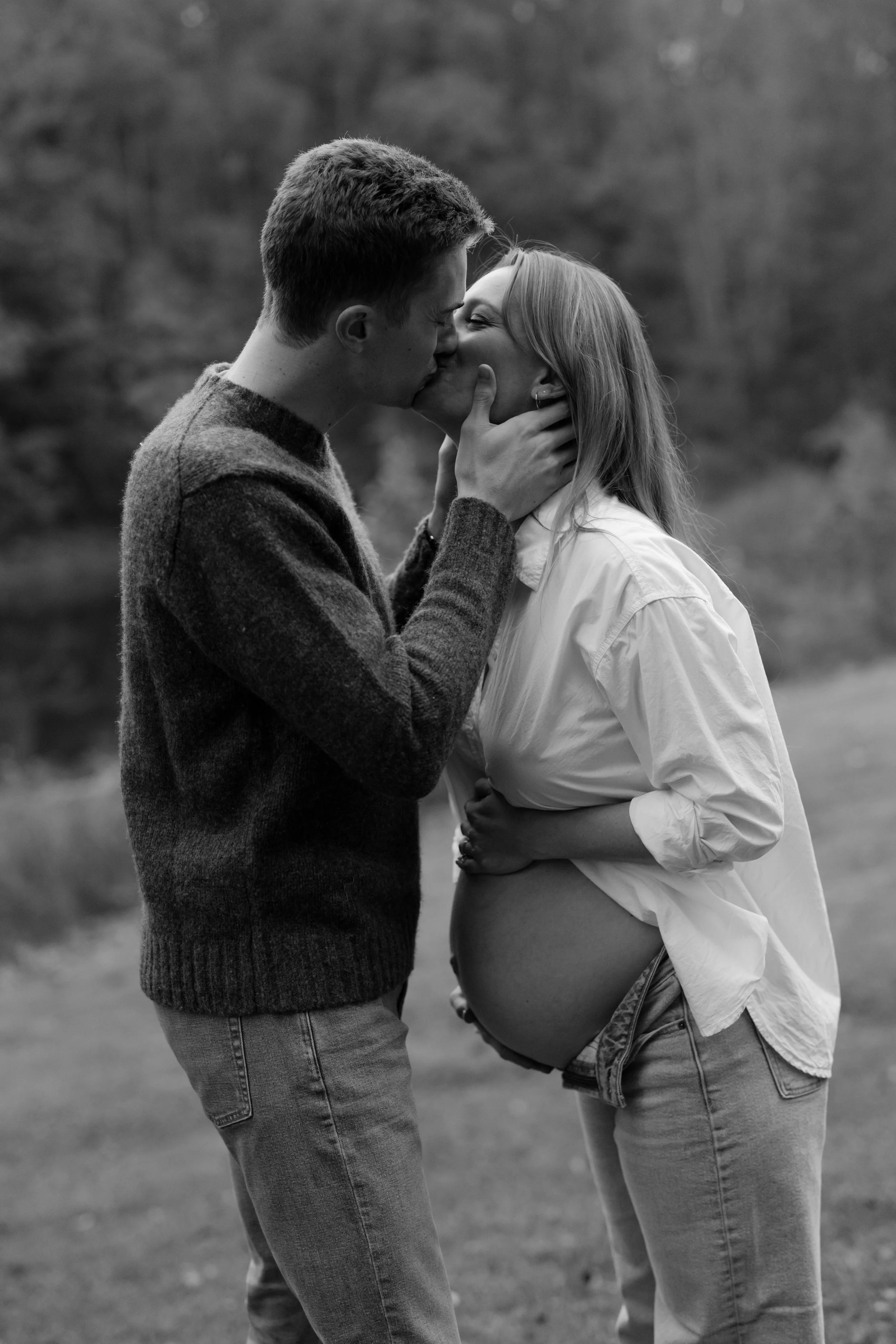 A couple kissing outdoors, with the woman pregnant, in a natural setting with trees in the background.