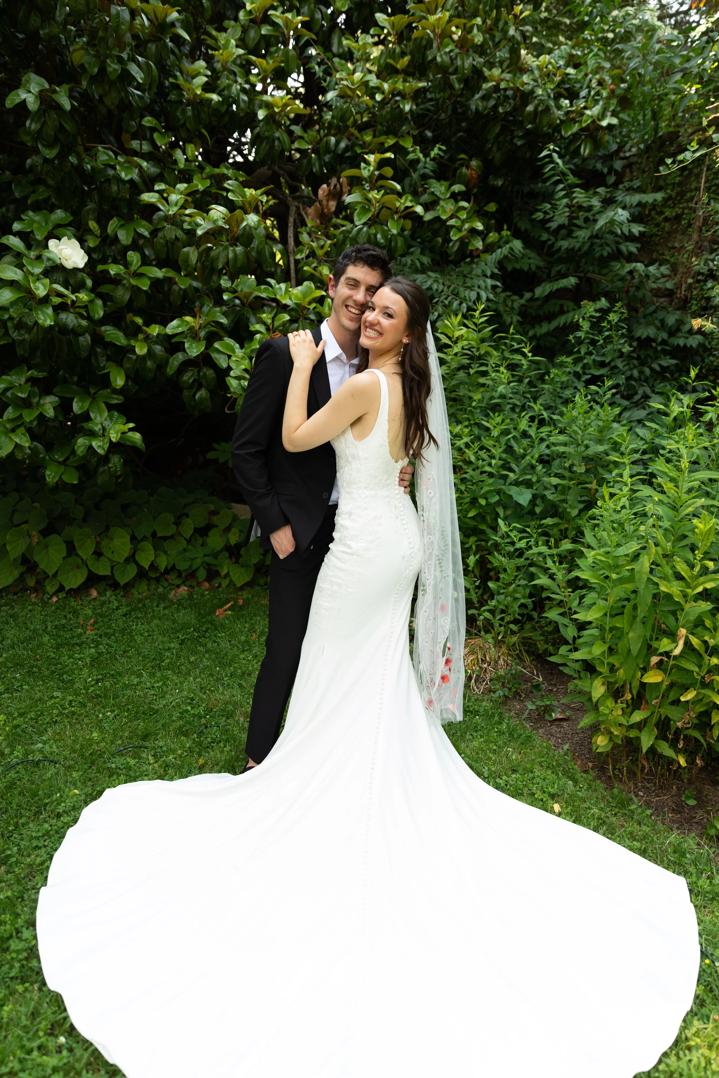 Happy bride and groom smiling, embracing outdoors in a garden setting, bride in a sleek white wedding dress with a long train and veil, groom in a black suit and white shirt.