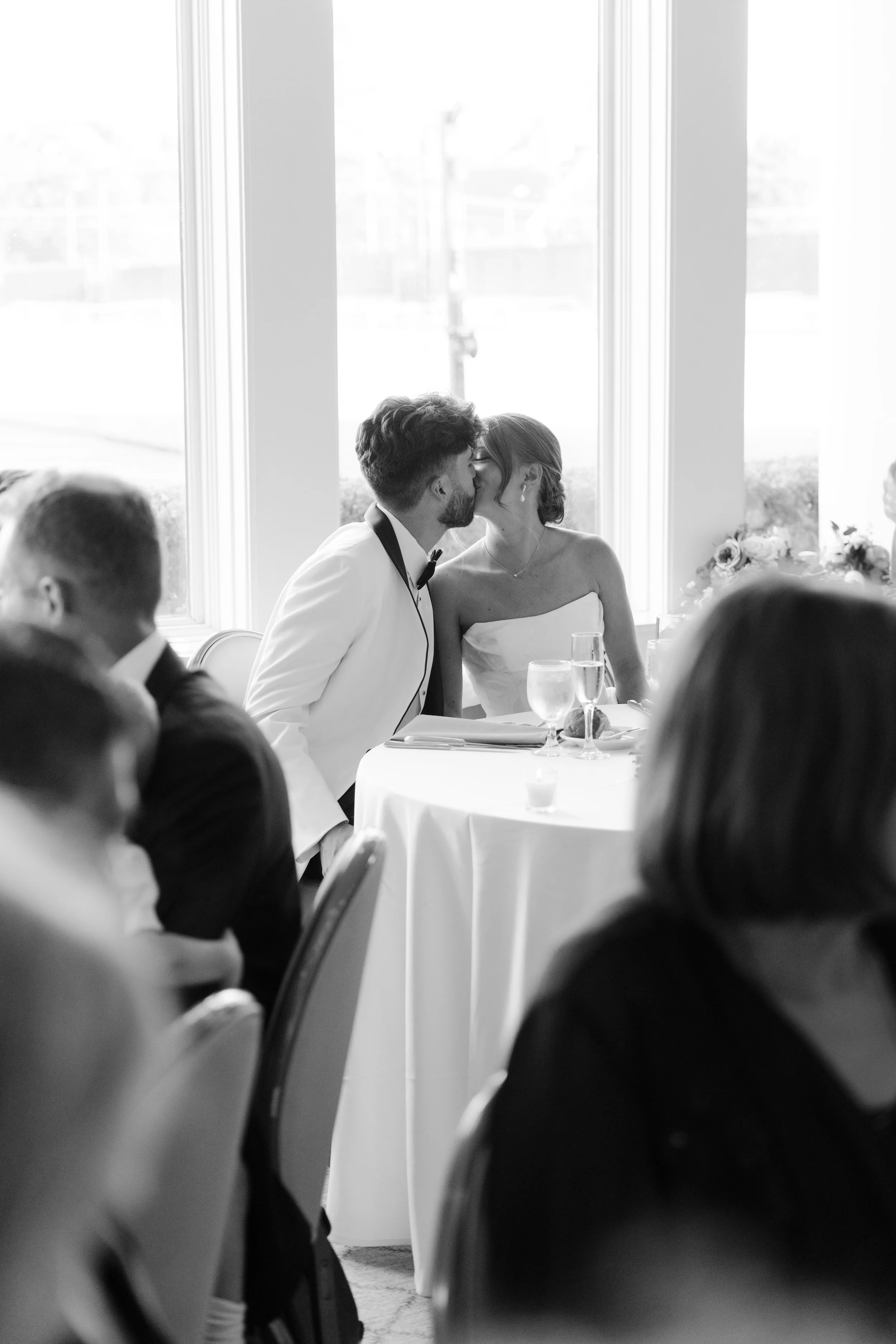 Black and white photo of a bride and groom sharing a kiss at their wedding reception, with guests seated at tables around them.