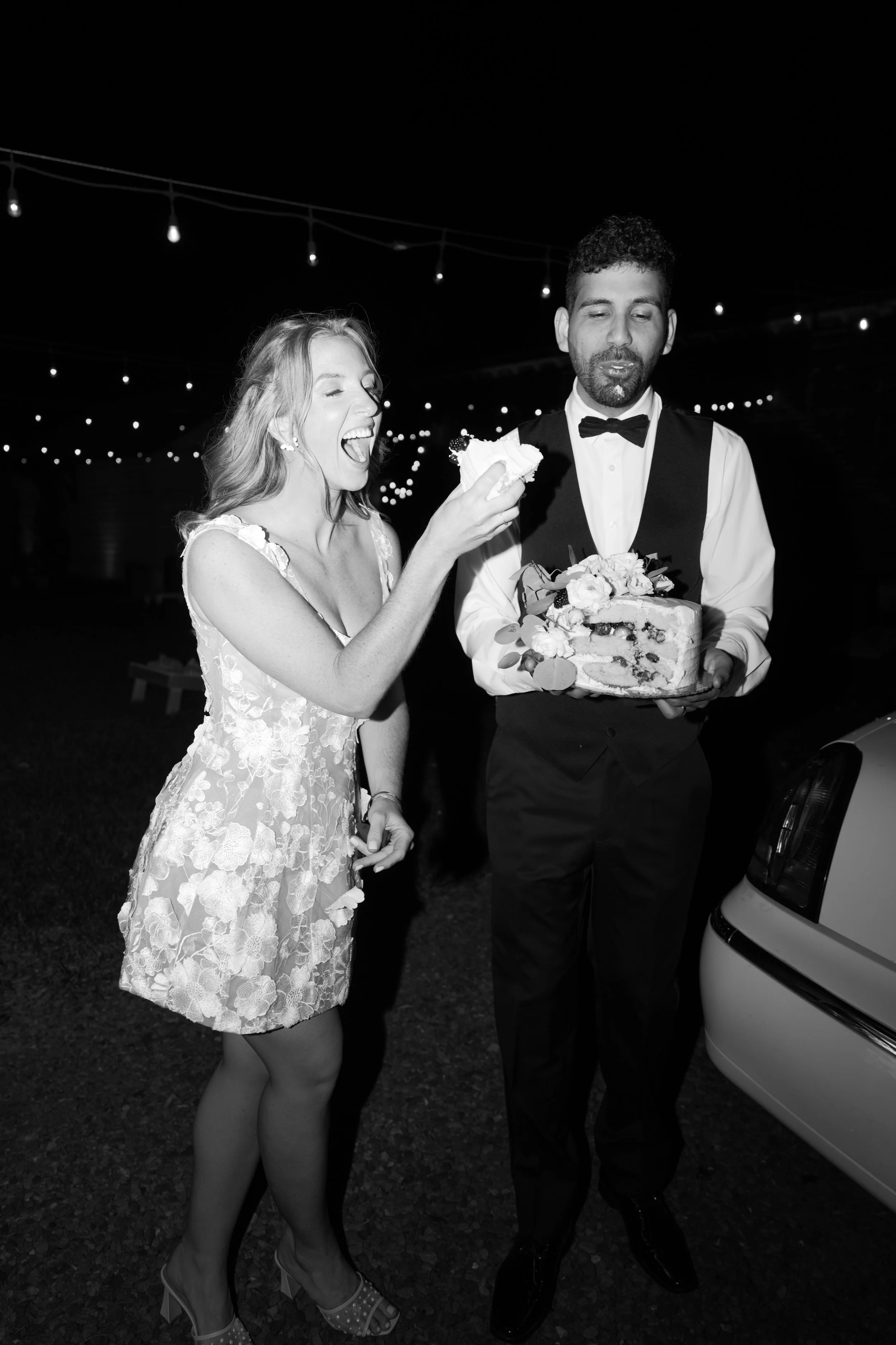 A woman in a floral dress and a man in a tuxedo celebrate at a nighttime outdoor event, with string lights above. The woman is holding a piece of cake and the man is holding a cake with flowers.